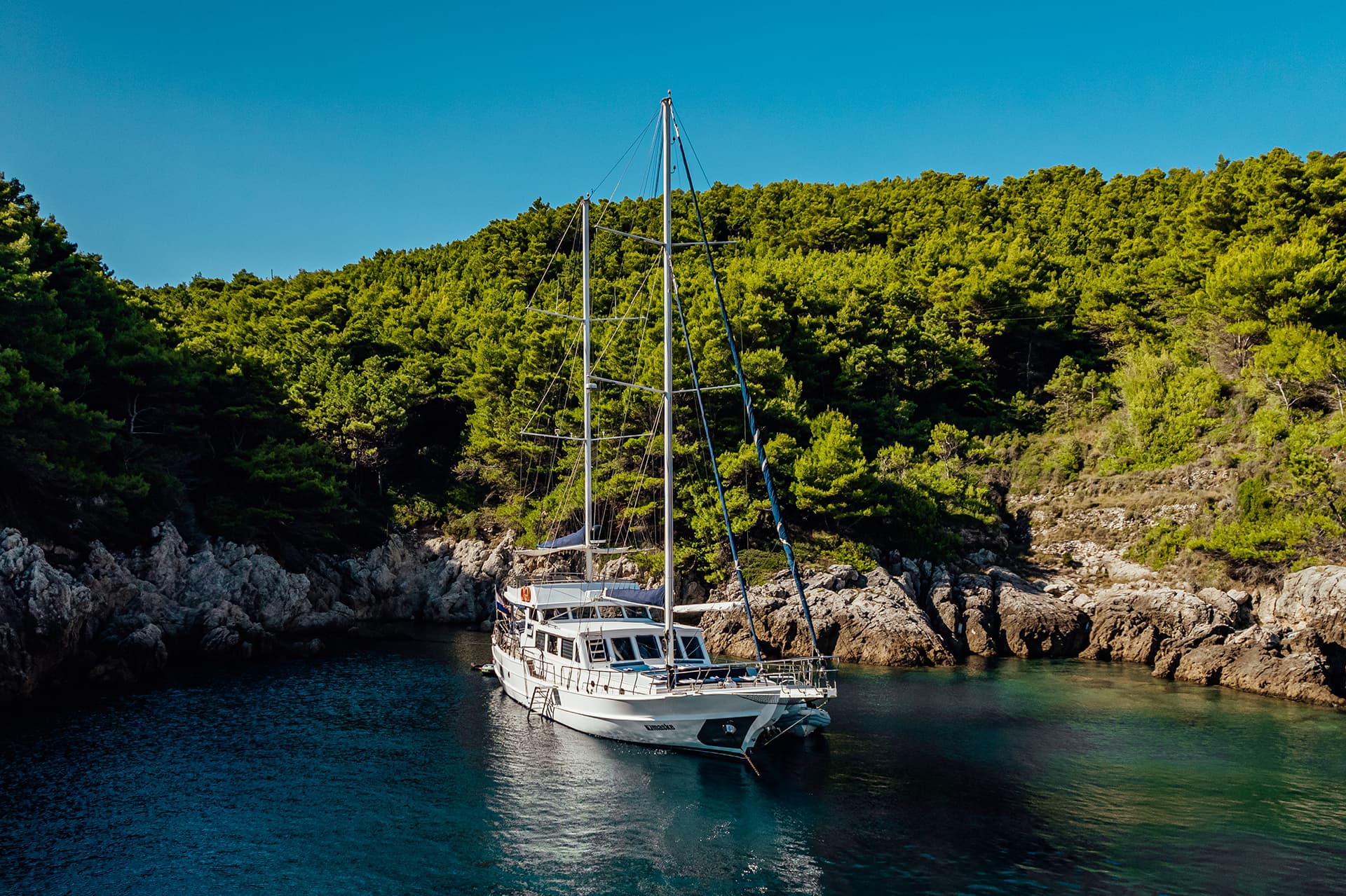 Gulet yacht named 'Maske' anchored in a cove with rocky shores and dense green trees.