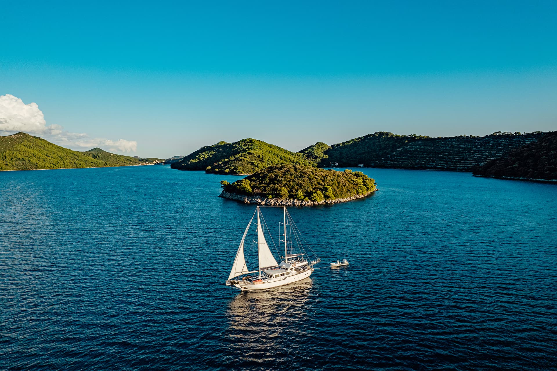 Sailboat with white sails on deep blue water near lush green islands under clear sky.