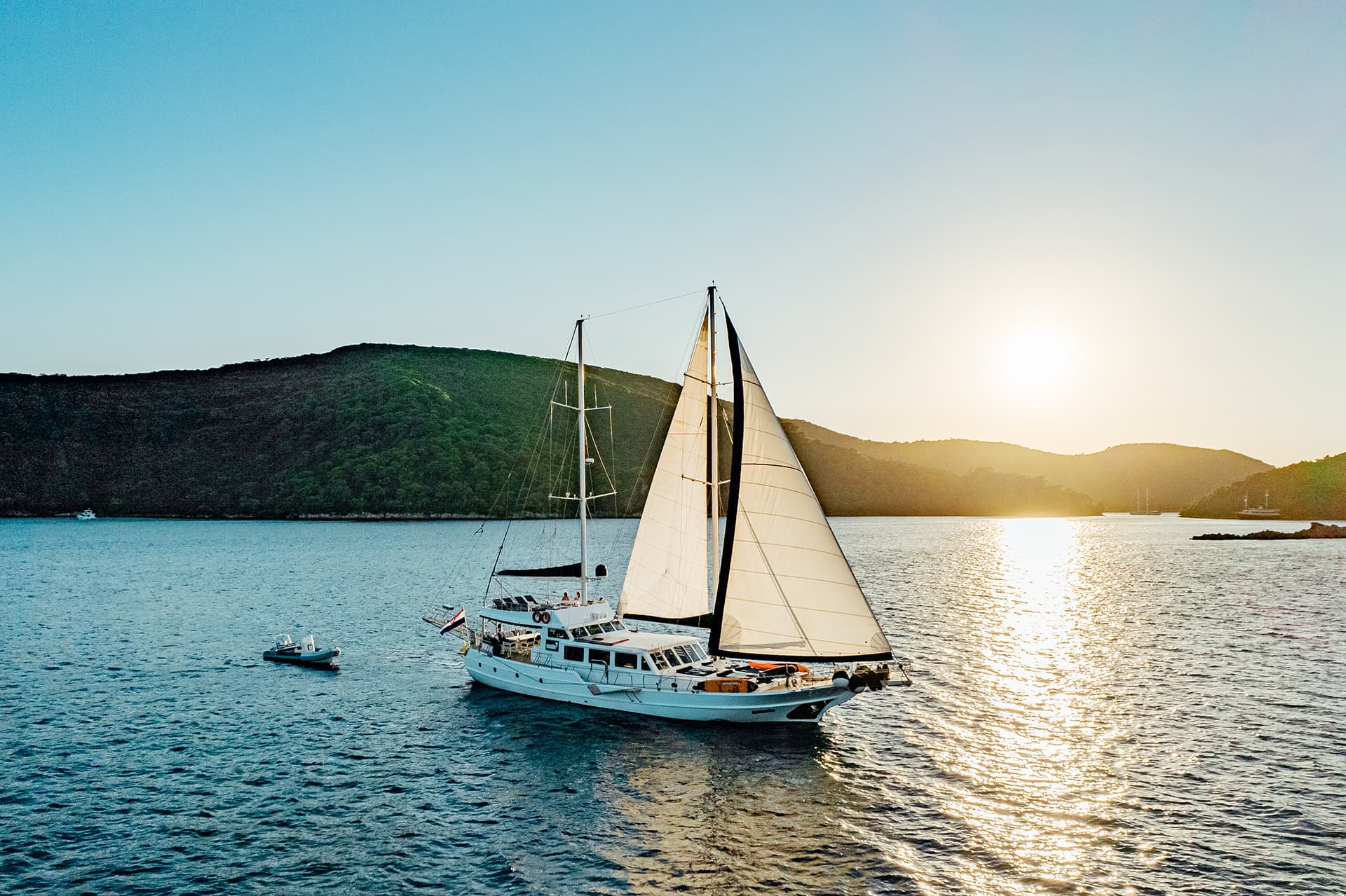 Gulet sailing boat with white sails on blue water near green hills at sunset