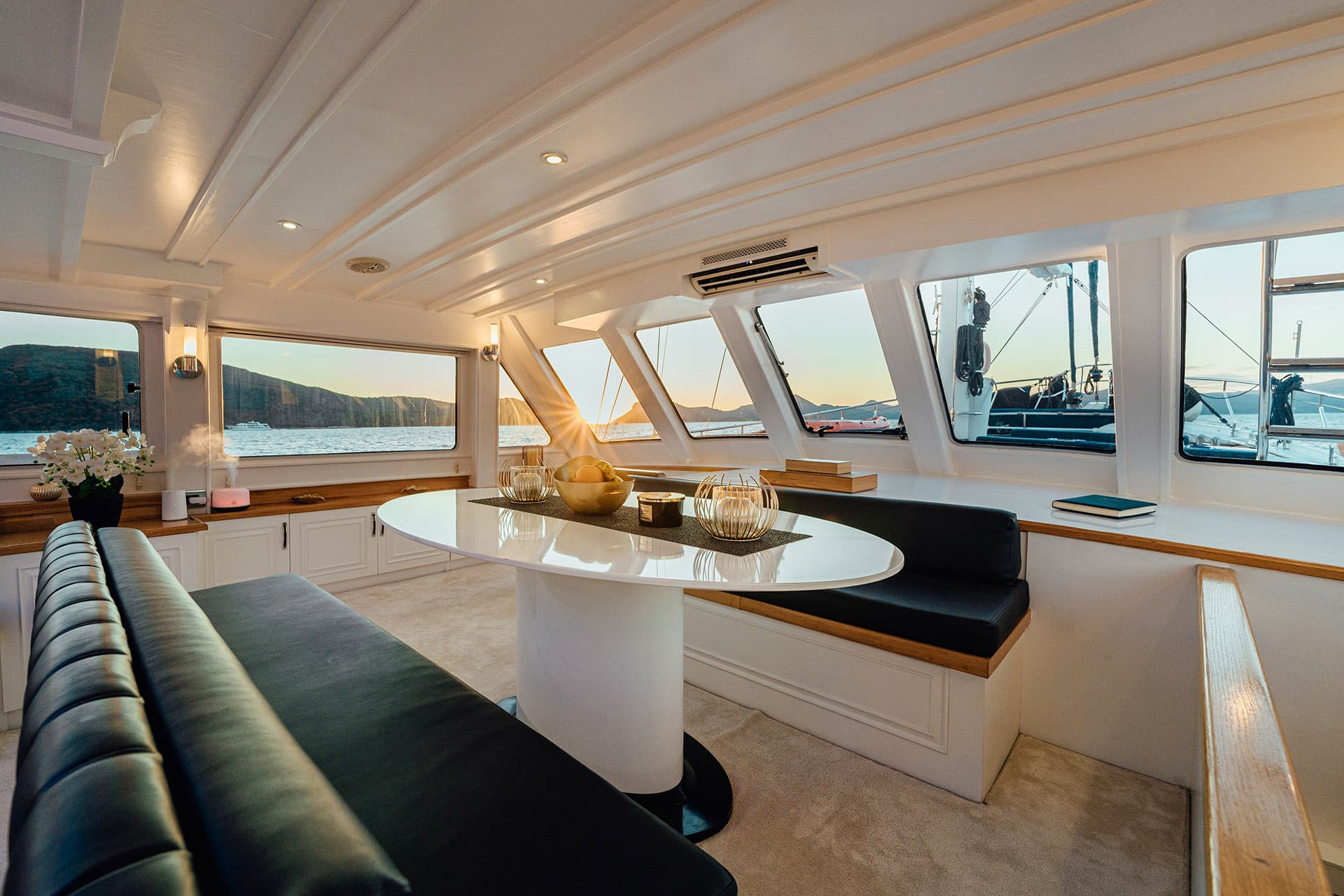 Interior dining area of a boat with white paneling and view of mountains and sea at sunset.