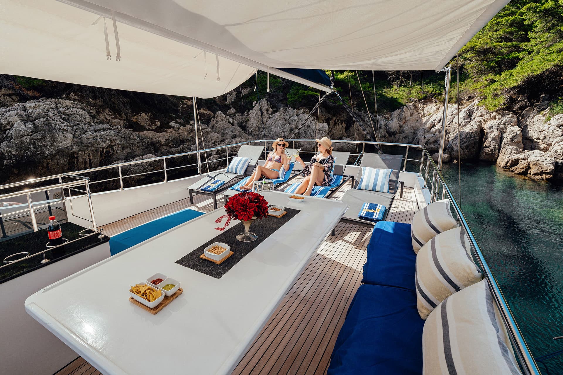 Two women sunbathing on deck of a gulet yacht near rocky, forested coastline.