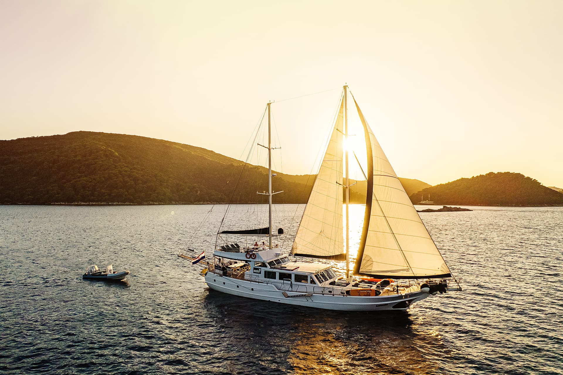 Gulet sailboat with sails up on the sea near green hills at sunset