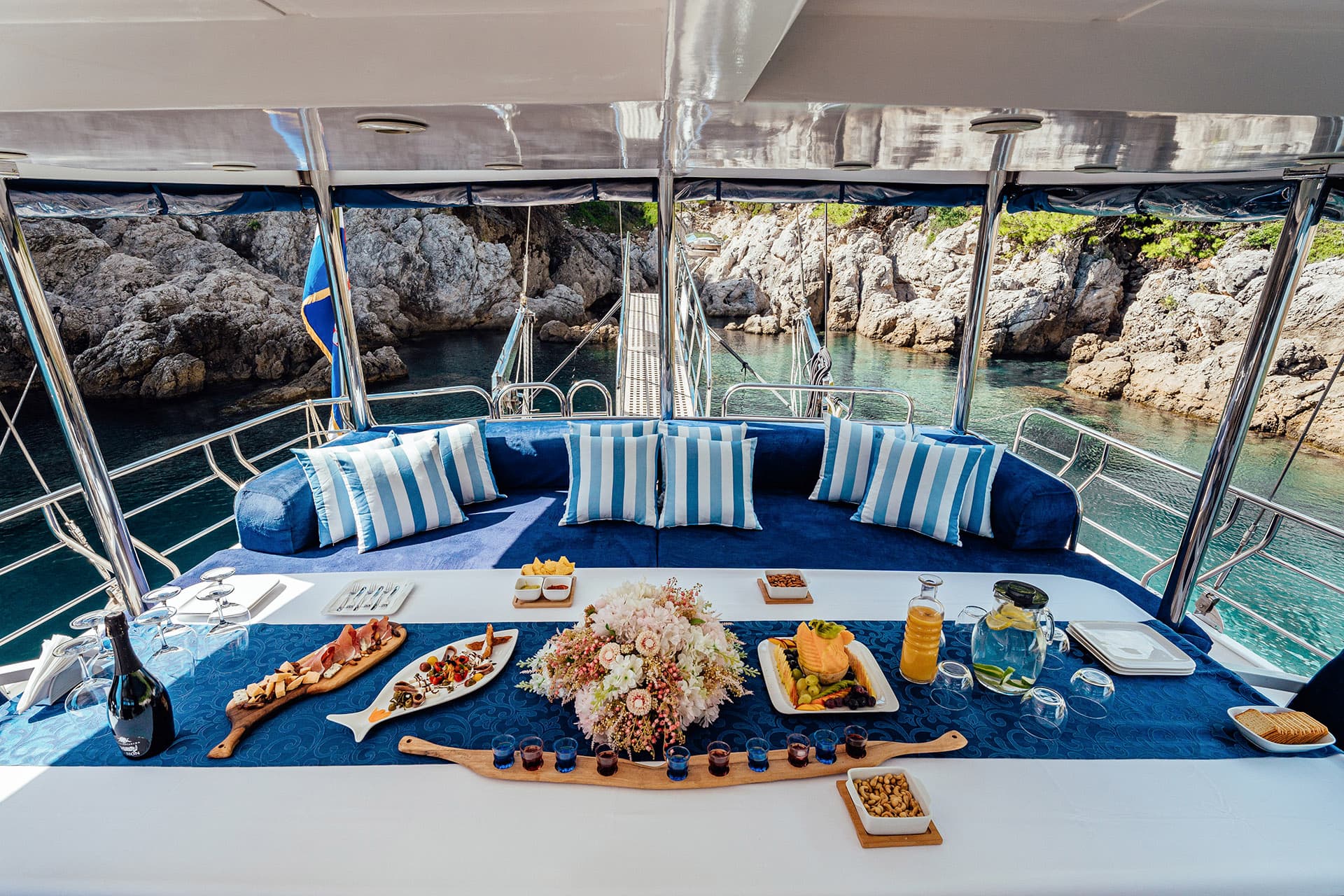 Al fresco dining on a yacht deck with appetizers, drinks, and flowers near rocky coastline.