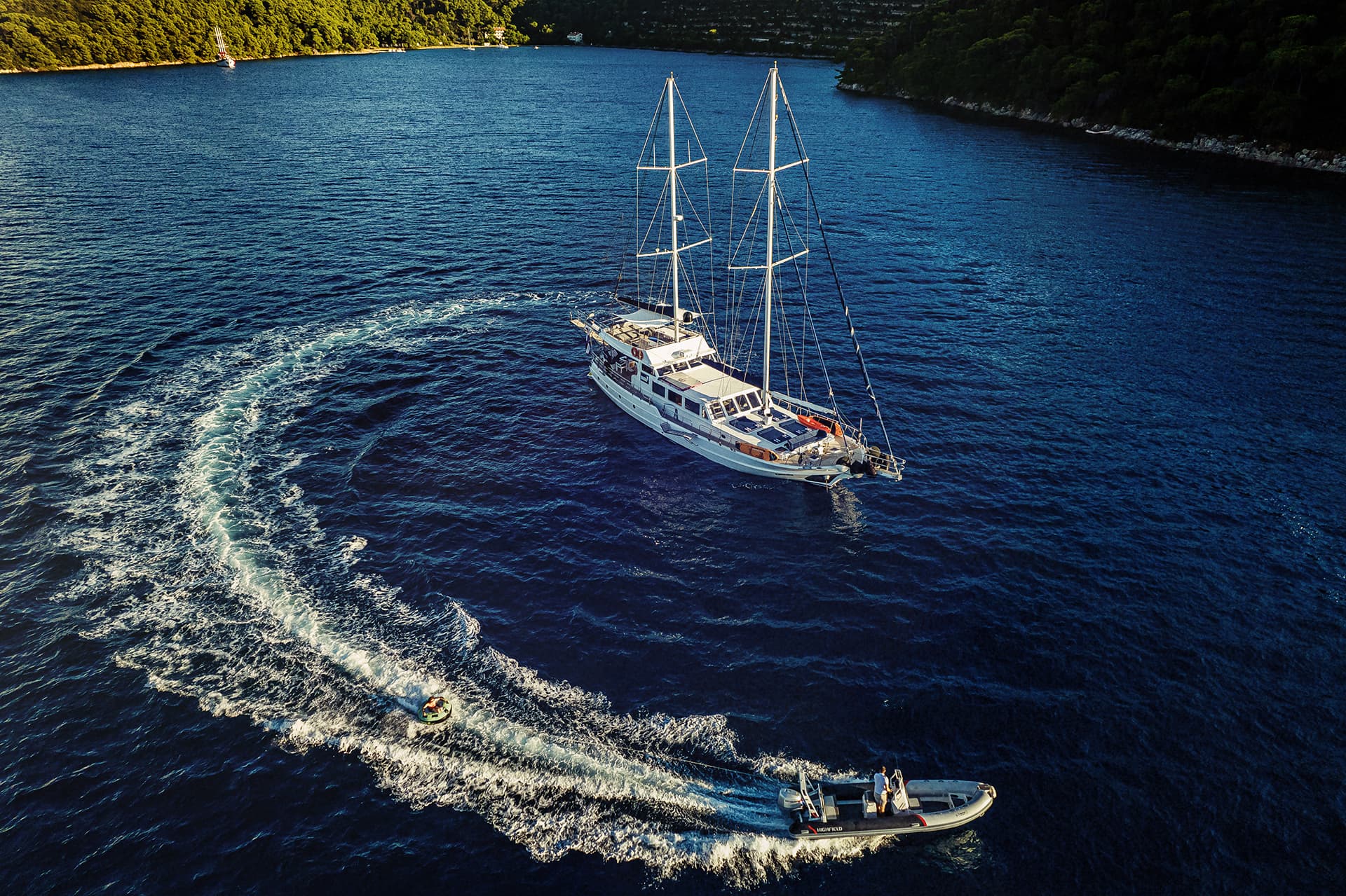 Gulet yacht anchored near forested coastline while dinghy tows inflatable ring on dark blue sea.