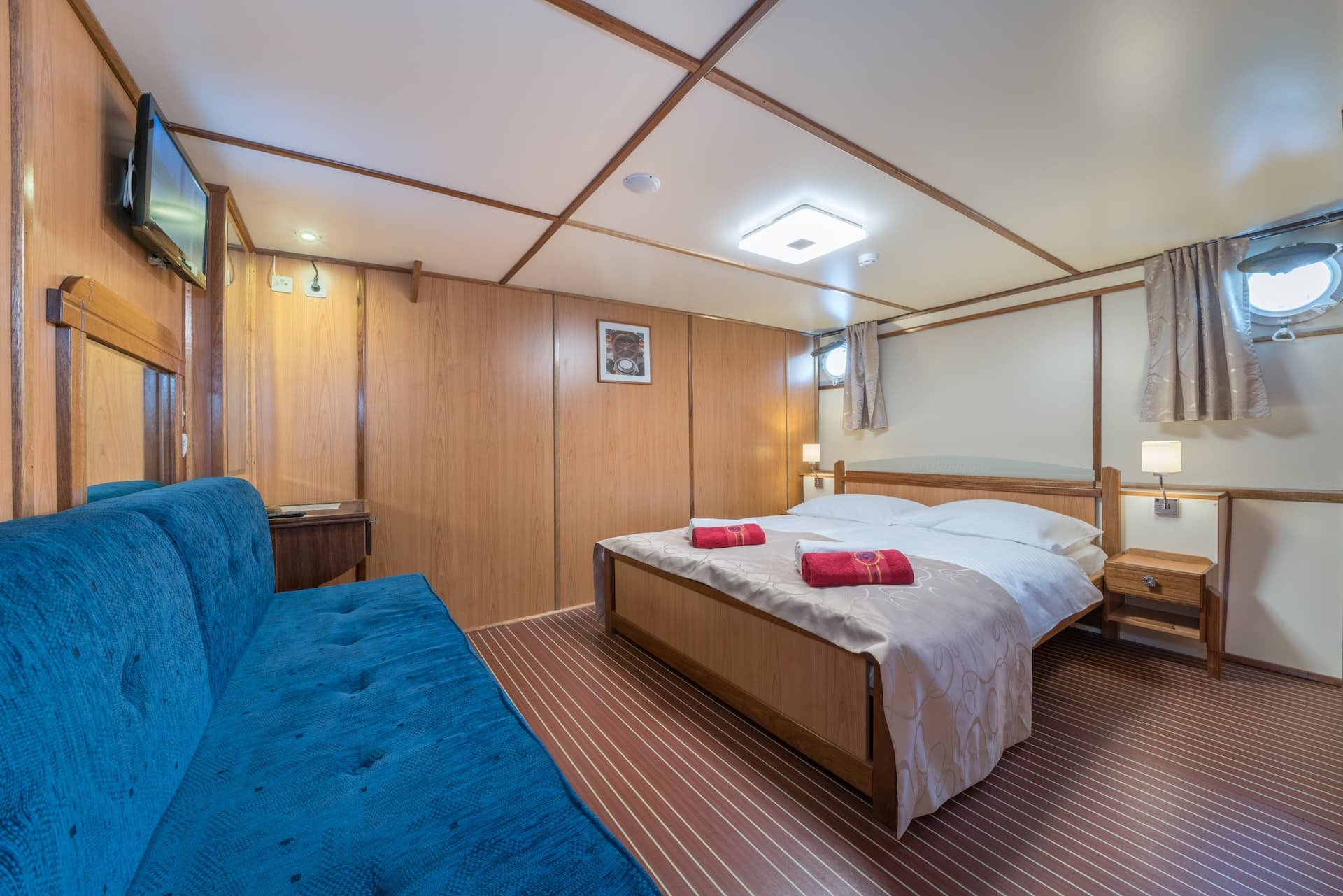Cabin interior with double bed, blue sofa, wood paneling, and porthole windows.
