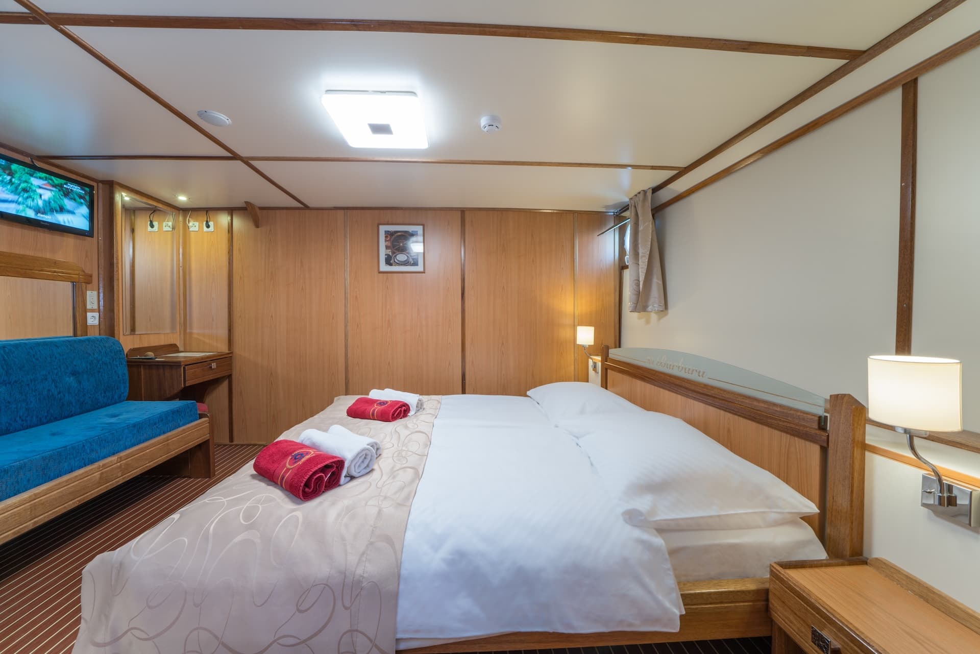 Cabin interior with double bed, blue sofa, wood paneling, and TV screen, likely on a boat.