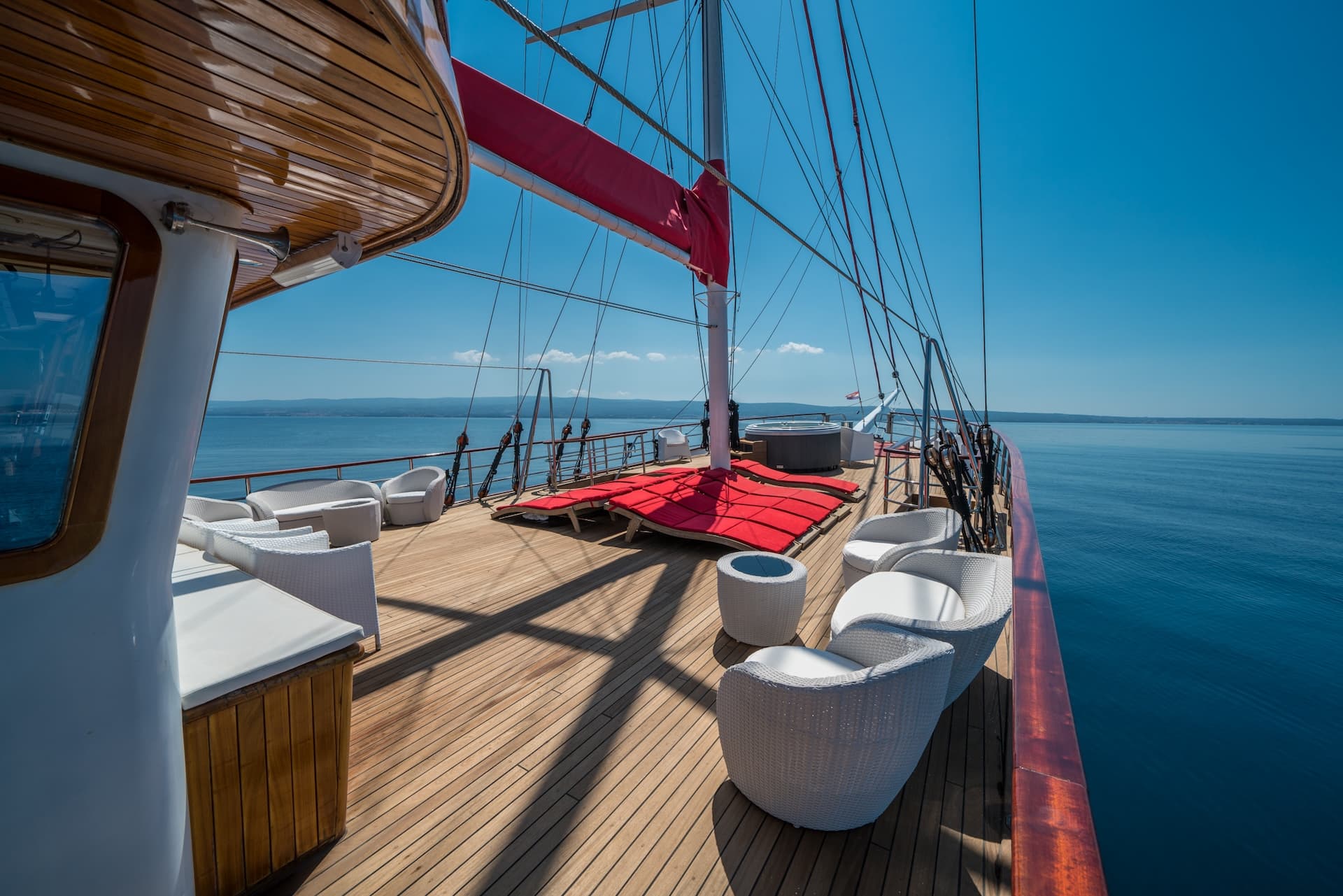 Deck of a large sailboat with white wicker furniture and red cushions on blue sea.