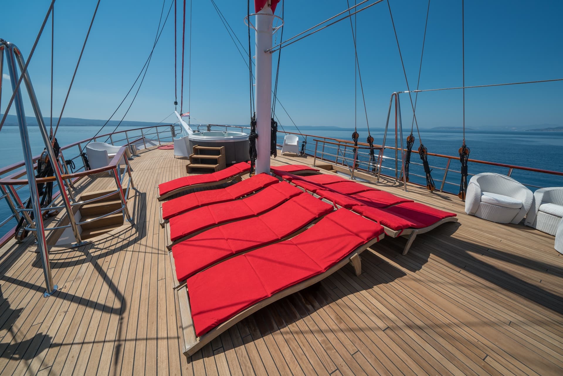 Sun deck with red loungers, hot tub, and white wicker chairs on a boat at sea.