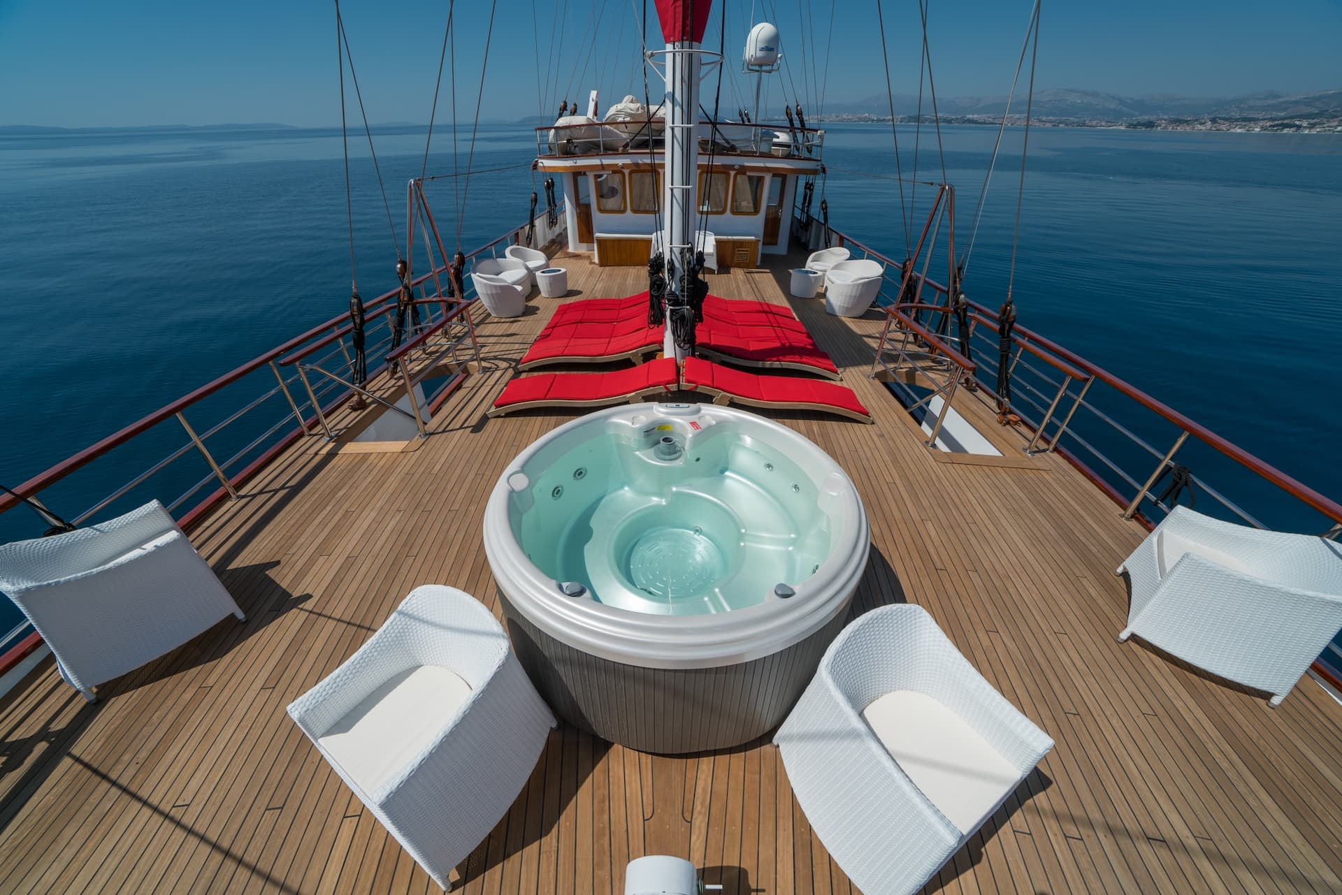 Hot tub and white wicker chairs on teak deck of boat sailing near coastline.