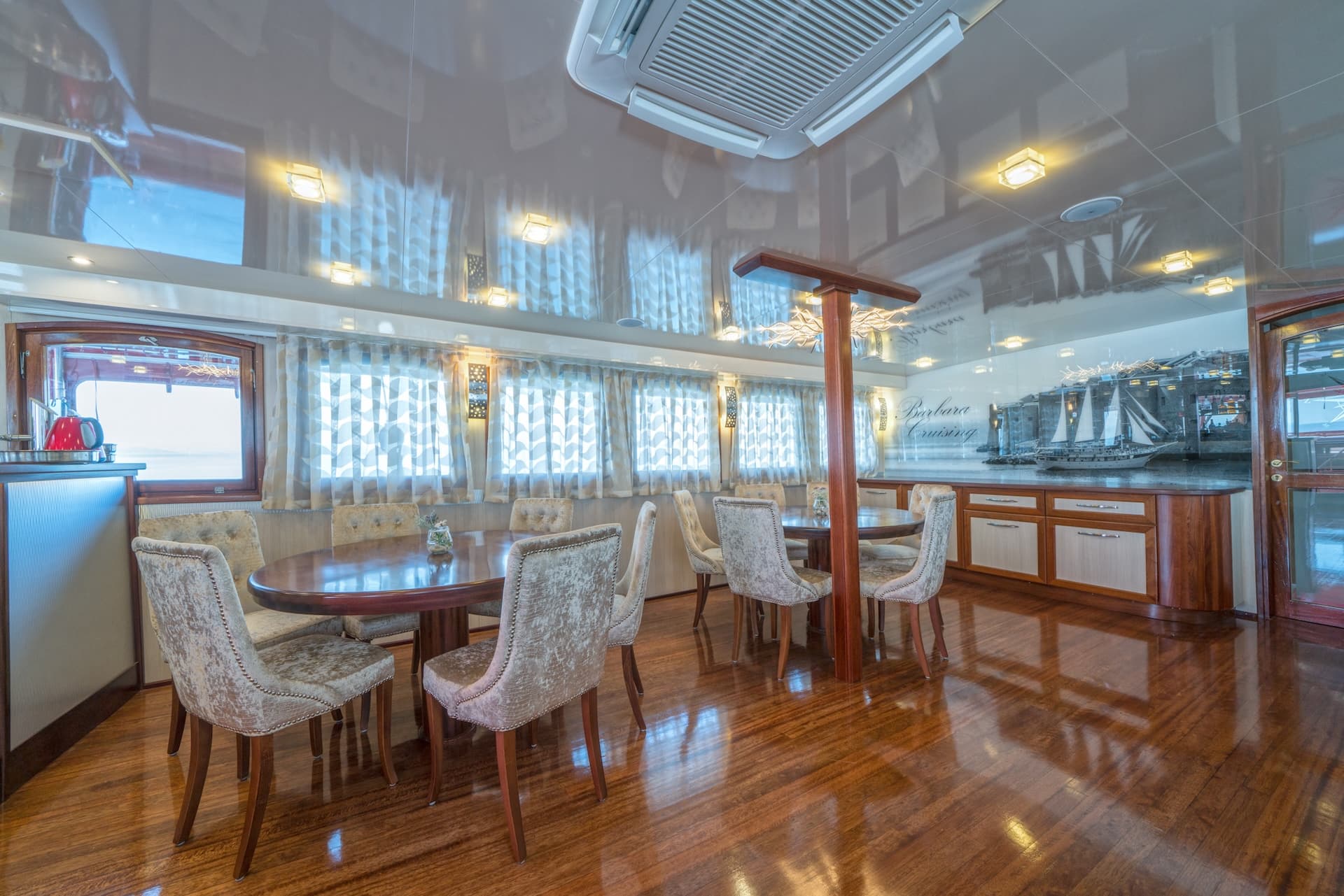 Dining area inside a boat with polished wood floors, upholstered chairs, and a ship mural.