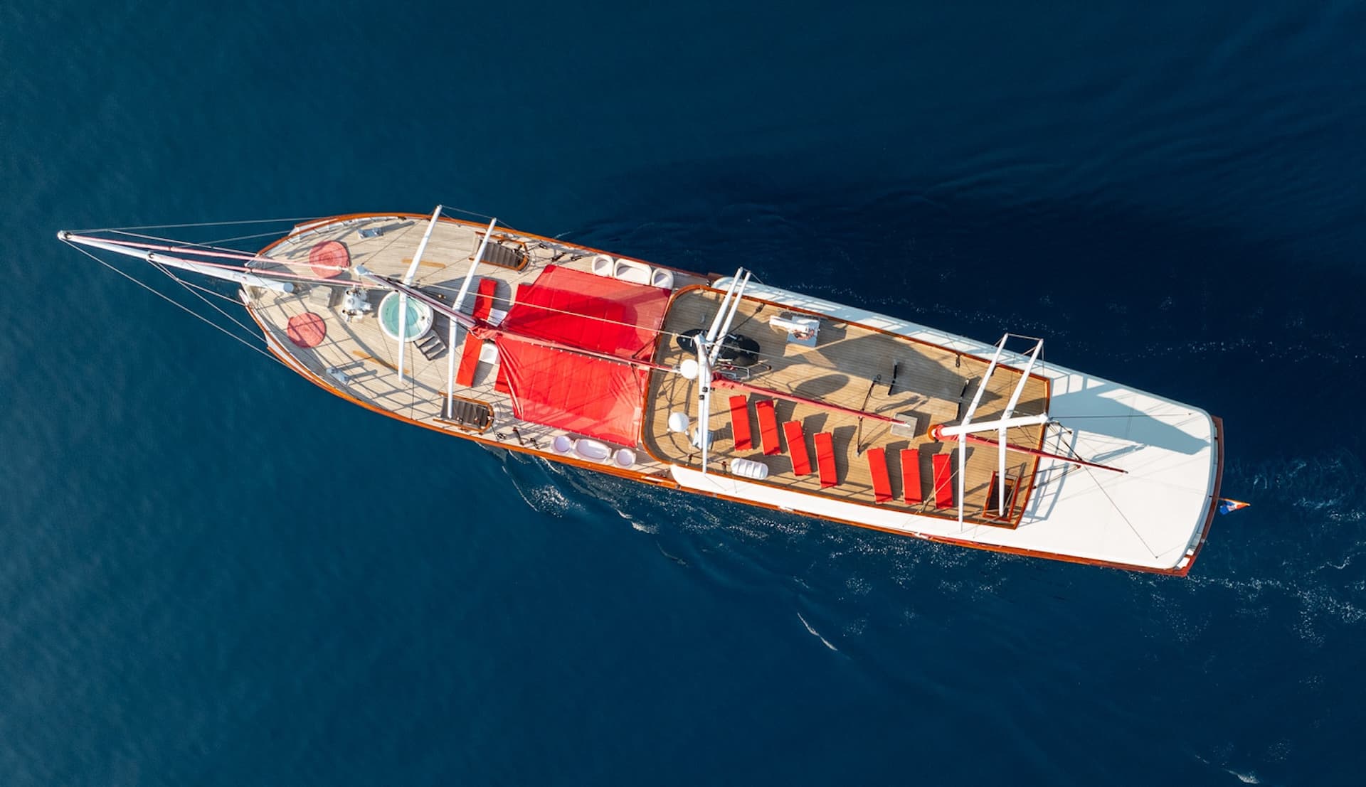 Aerial view of a wooden gulet boat with red sunbeds and a jacuzzi on deep blue sea water.