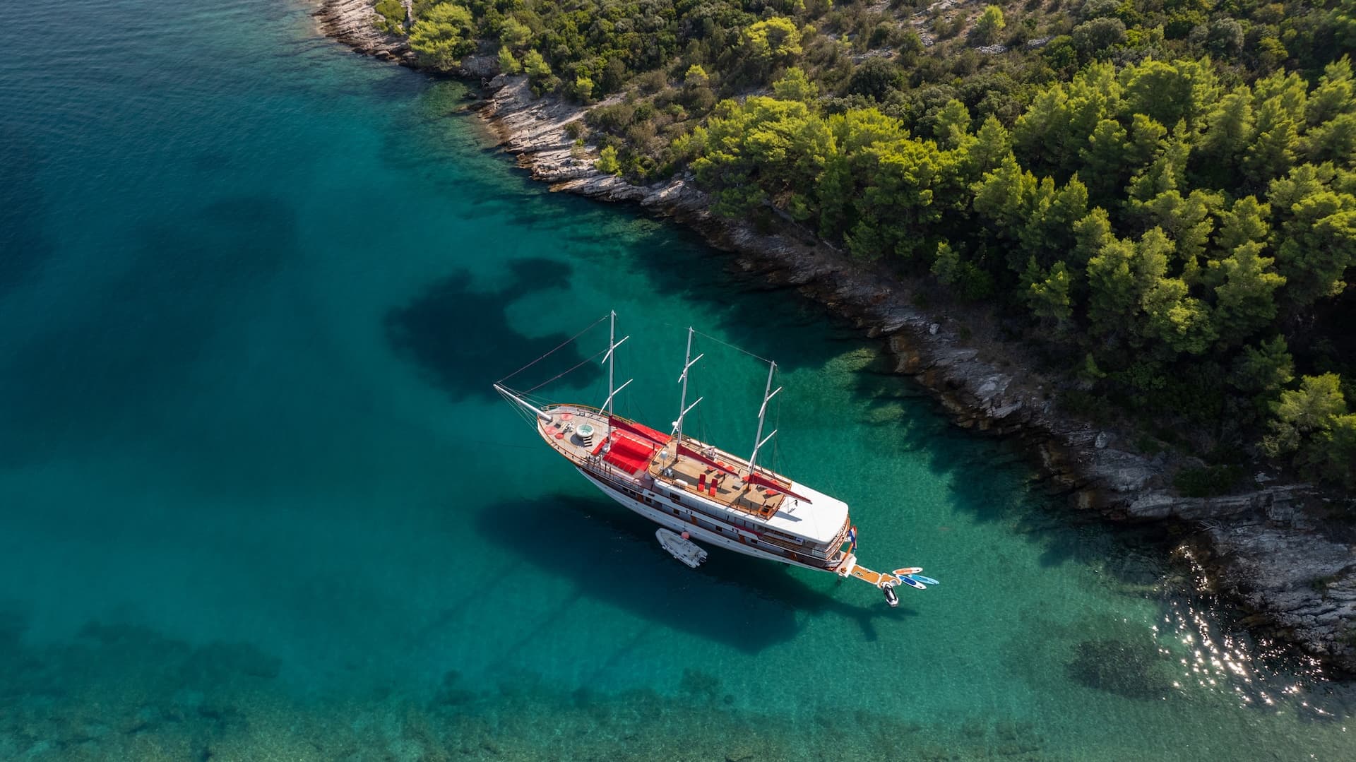 Gulet Barbara anchored in turquoise lagoon near forested rocky coastline.