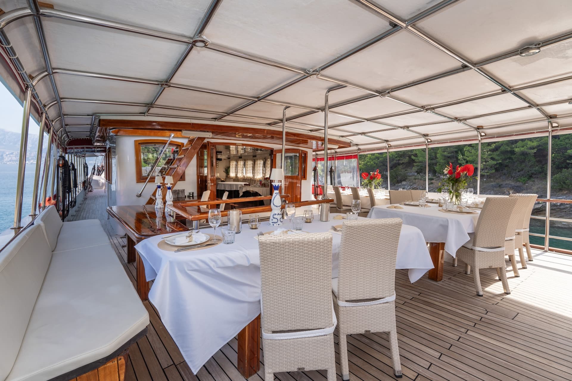 Dining area set on a gulet boat deck with white tablecloths near a wooded coastline.