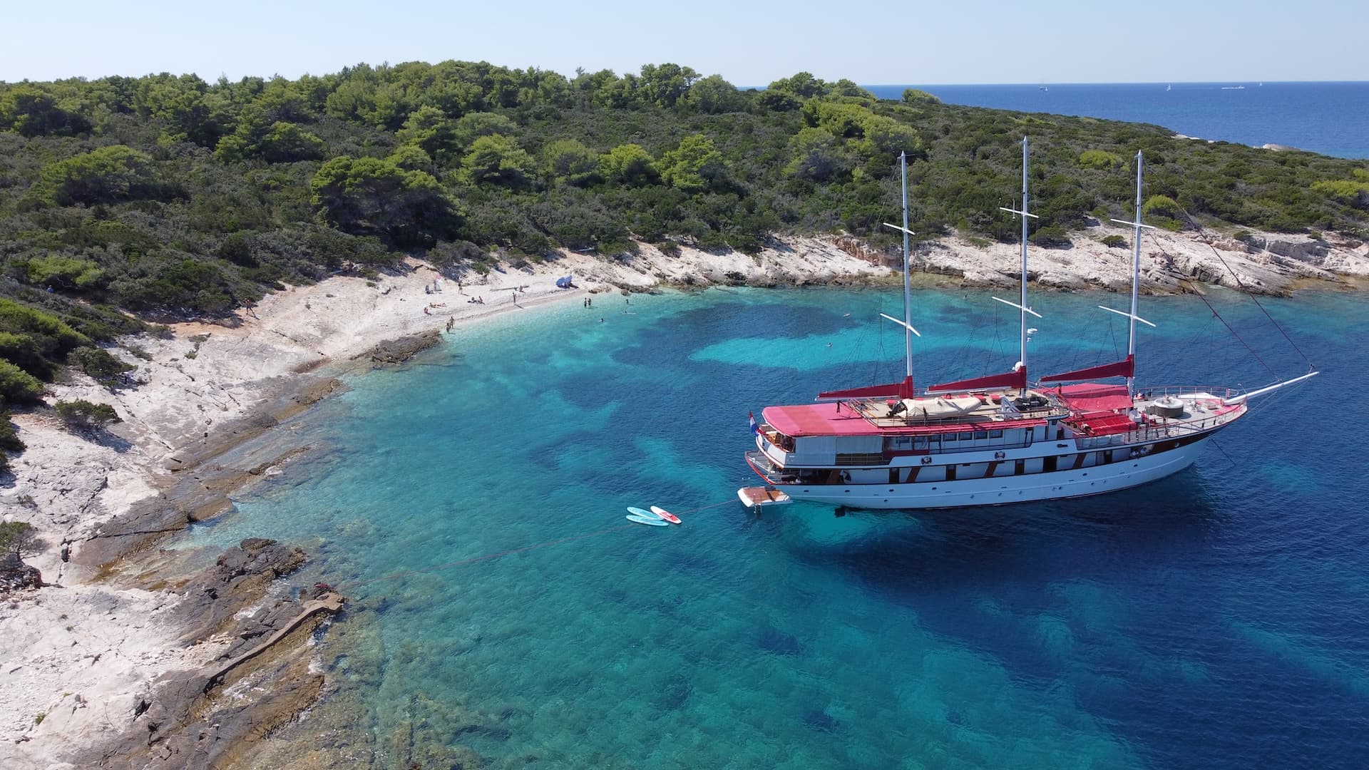 Gulet Barbara boat anchored in clear turquoise water near a rocky, tree-covered coastline.