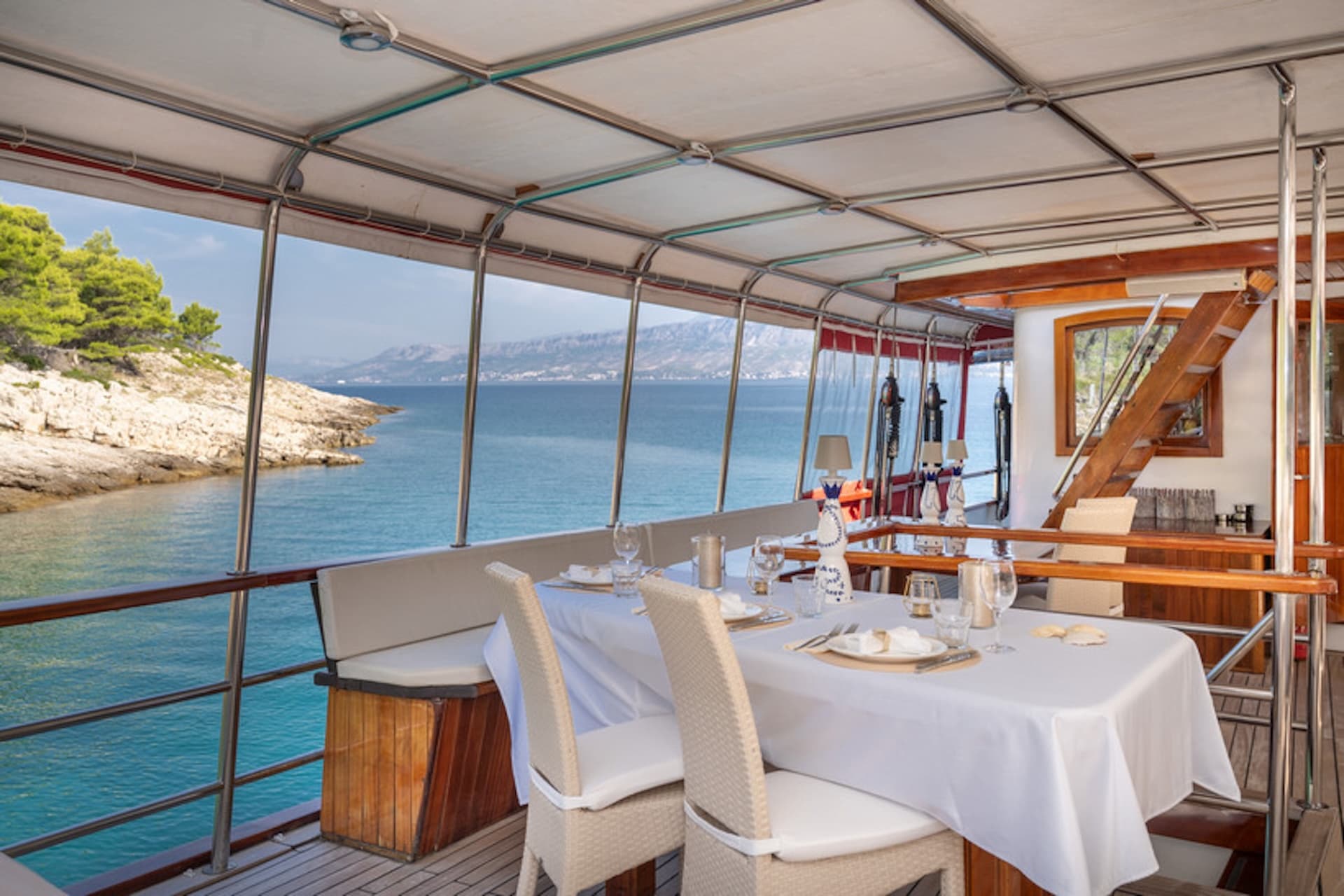 Outdoor dining table on a Gulet Barbara boat near a rocky, tree-lined coast.