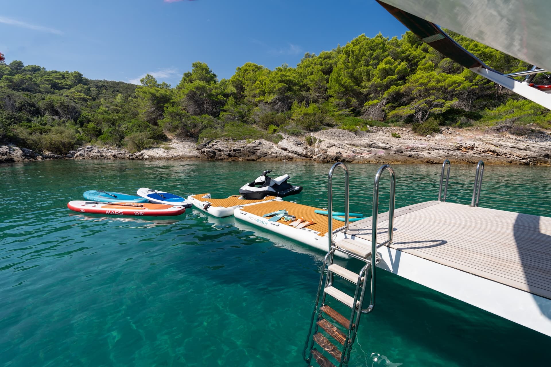 Water toys and swim platform moored in clear turquoise water near a rocky, forested coastline.
