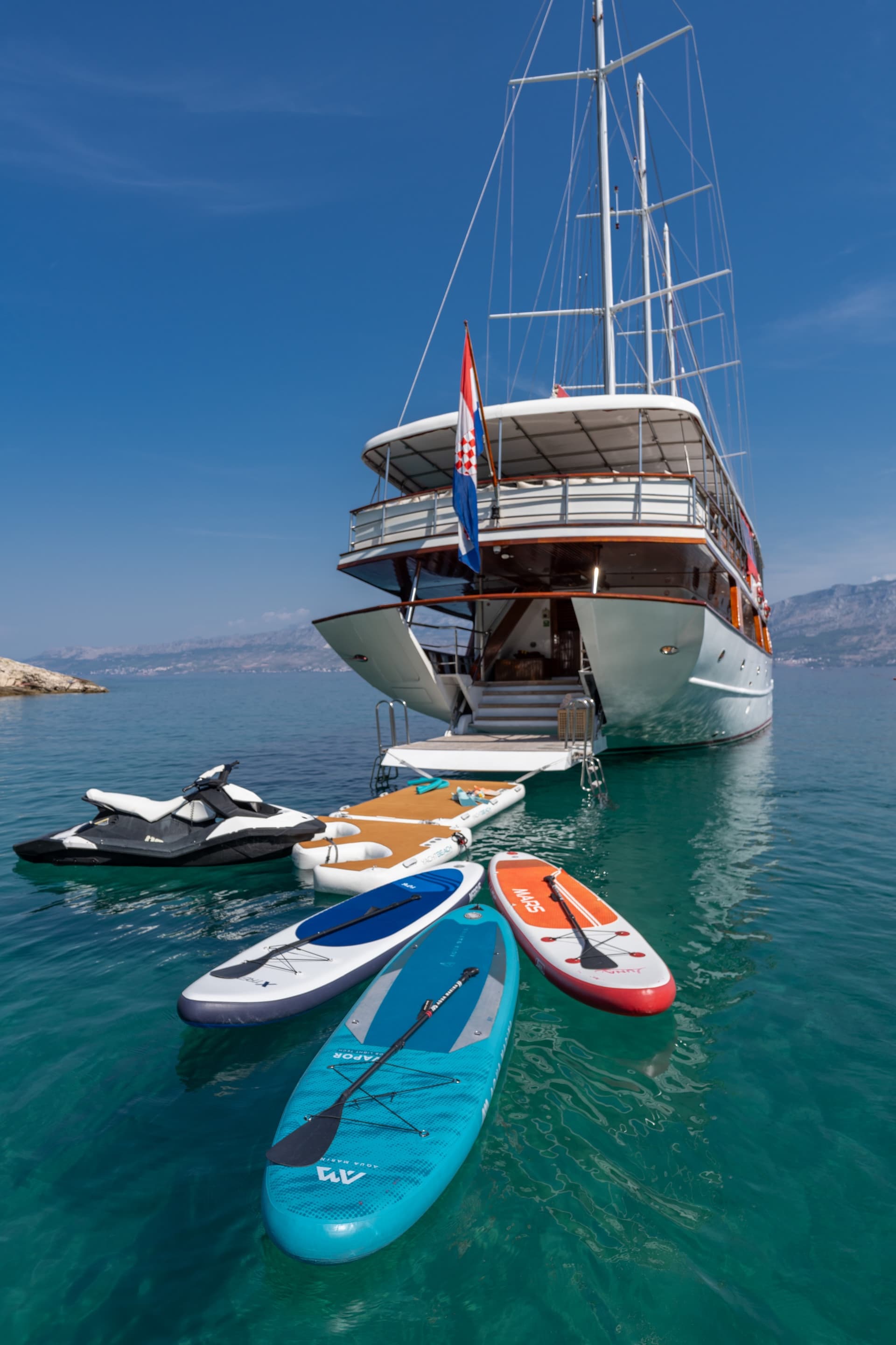 Gulet yacht with Croatian flag anchored near mountains, surrounded by paddleboards and a jet ski.