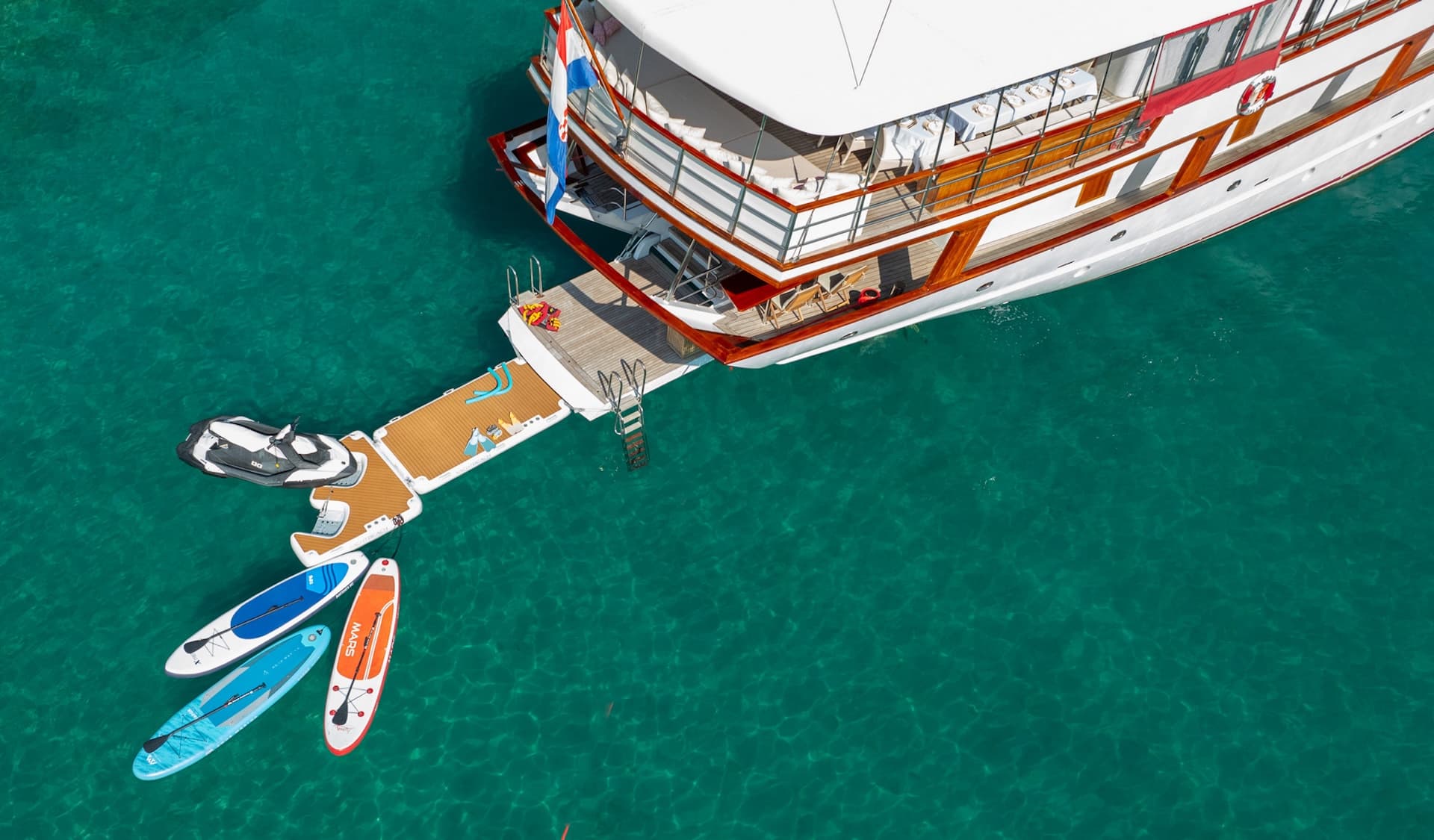 Gulet boat with water toys including jet ski and paddleboards anchored in clear turquoise water.