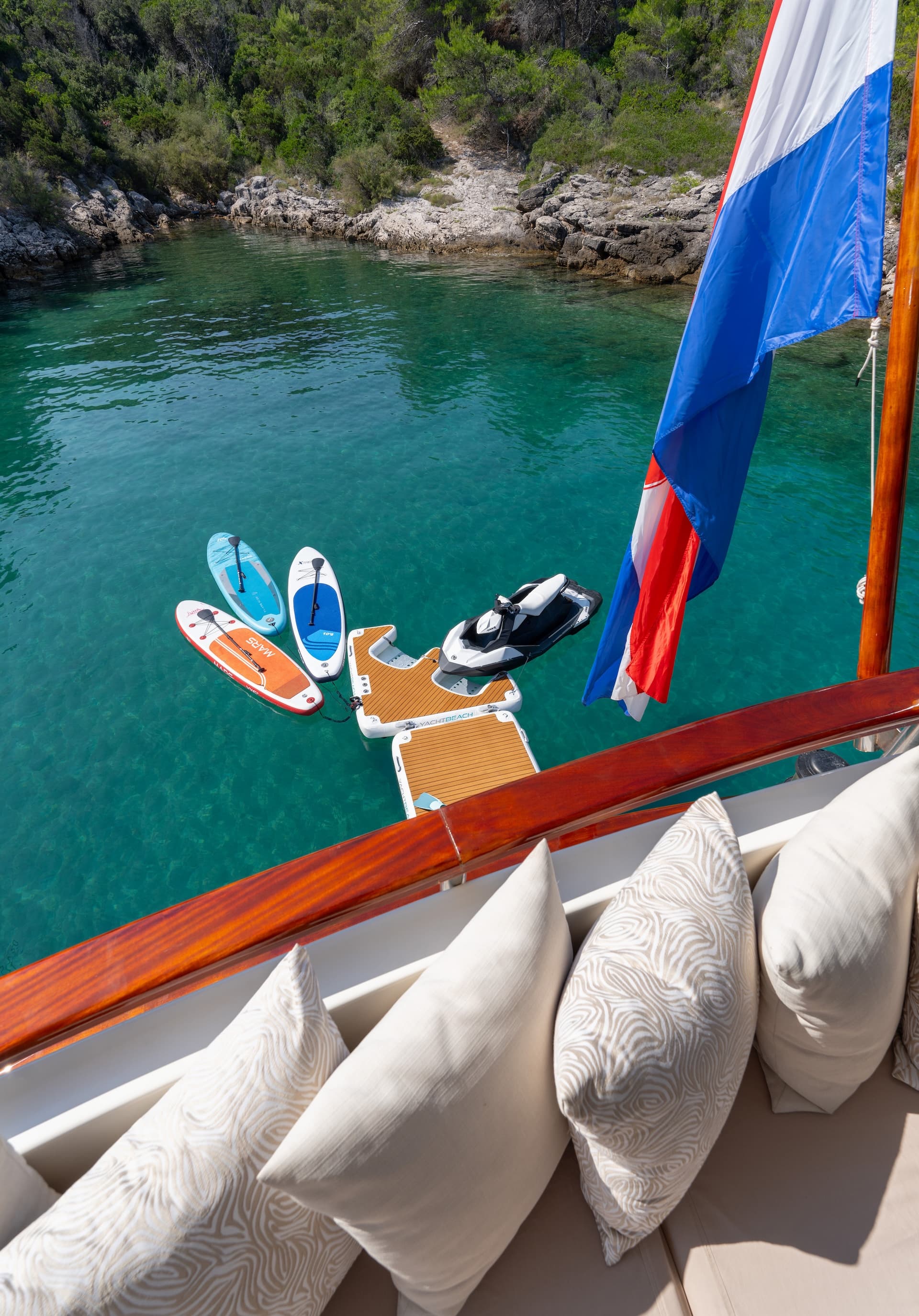 Water toys and jet ski floating near a gulet with a flag in clear turquoise cove water.