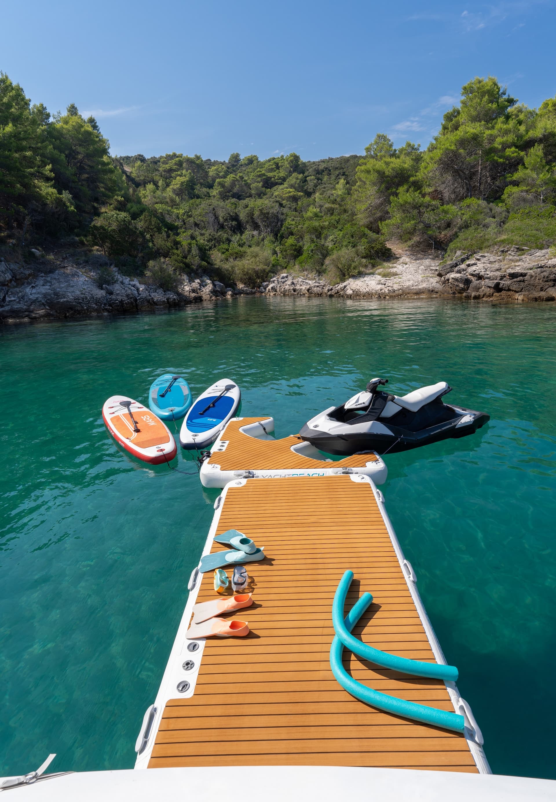 Inflatable swim platform with paddleboards and jet ski in clear turquoise cove near forested shore.