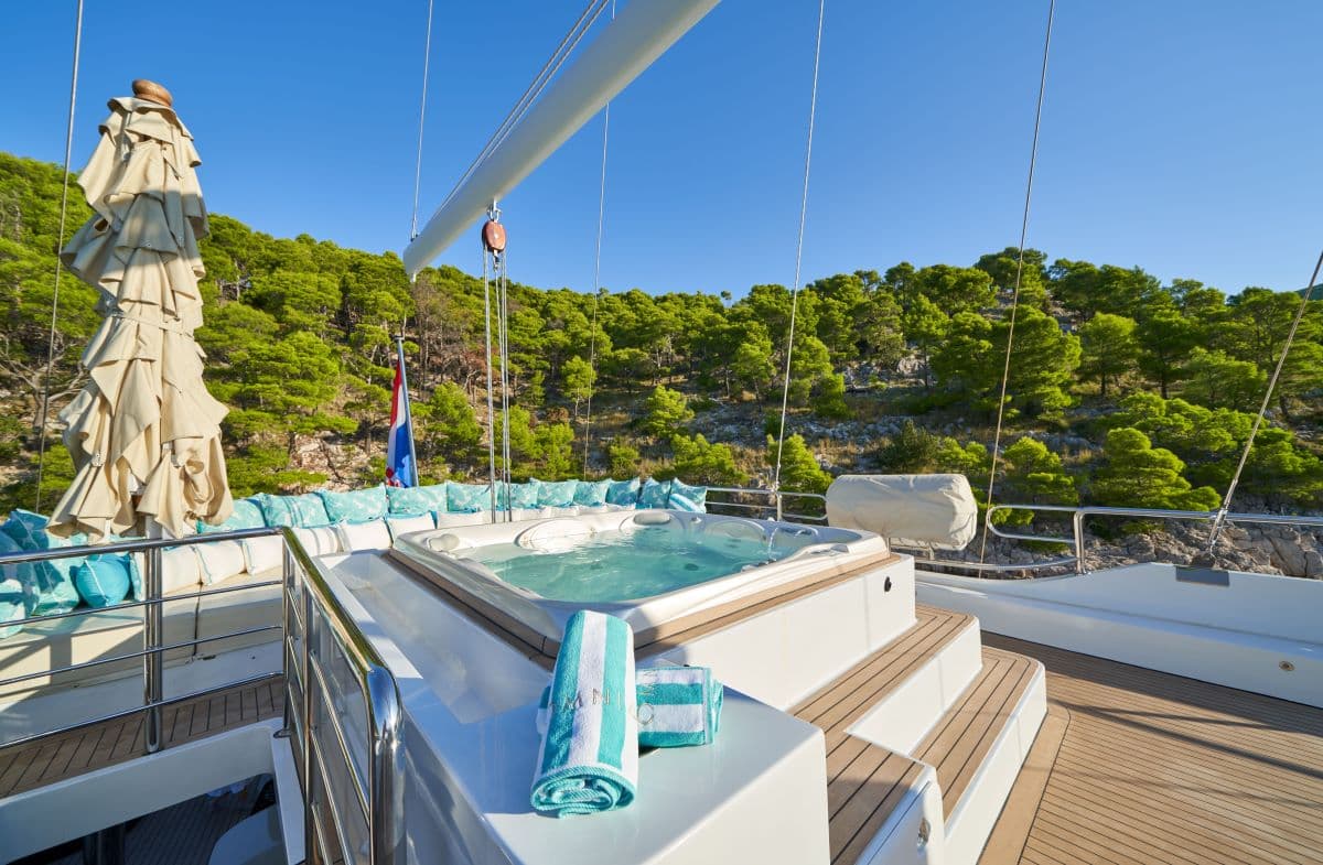 Jacuzzi on yacht deck with Croatian flag near lush green coastline under clear blue sky.