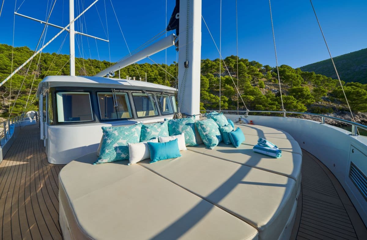Yacht sunbathing deck with turquoise pillows near wooded coastline under blue sky