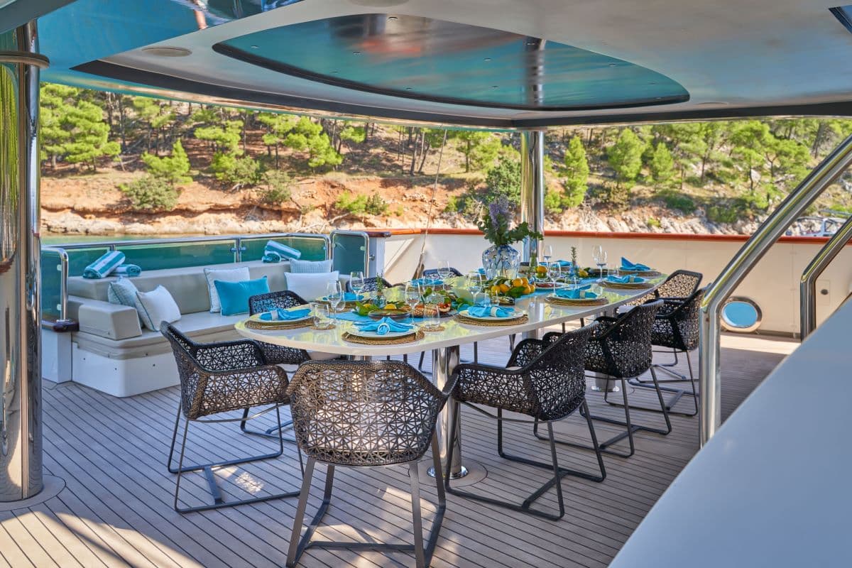 Outdoor dining table set for lunch on a yacht anchored near a wooded coastline.