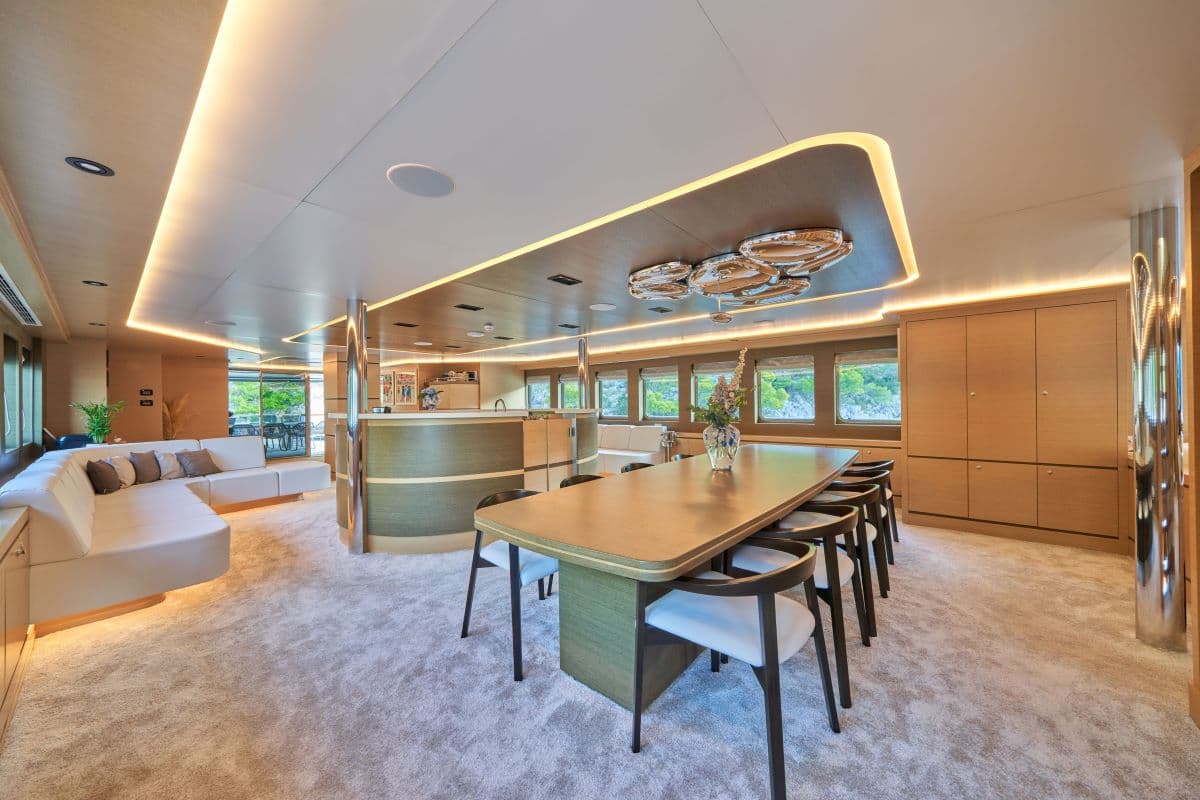 Modern yacht interior with dining table, lounge seating, and wood paneling overlooking green coastline.
