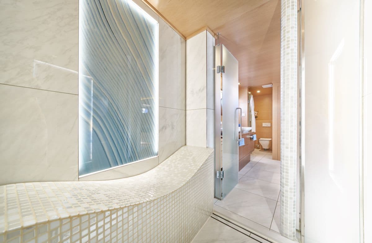 Modern spa area with backlit blue wave panel, white mosaic tile bench, and view into wooden bathroom.