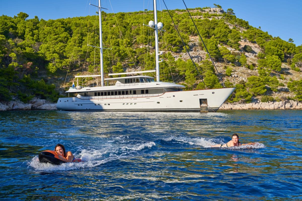 Two people playing on water toys near a large yacht anchored by a green, rocky coastline.