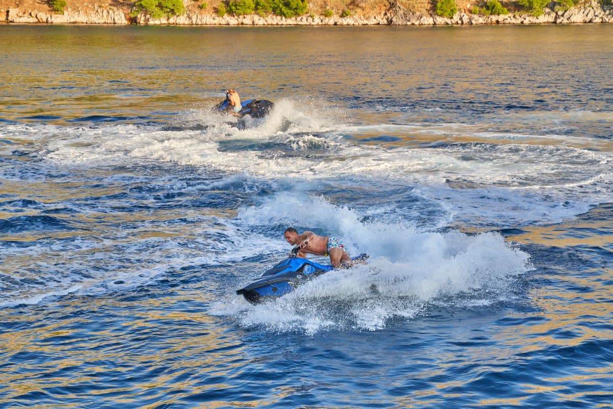 Two people jet skiing on blue water near a tree-lined rocky coastline.