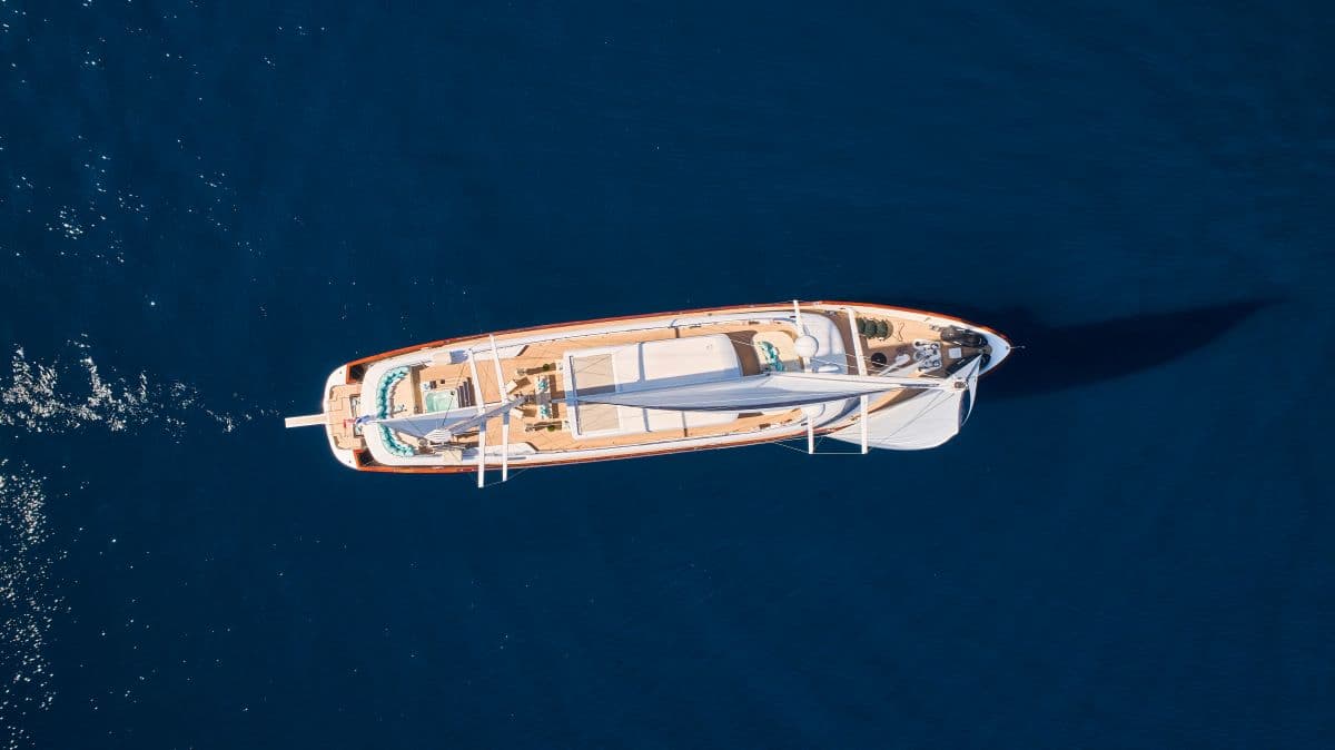 Luxury yacht with wooden deck and white sail on deep blue ocean water, aerial view.