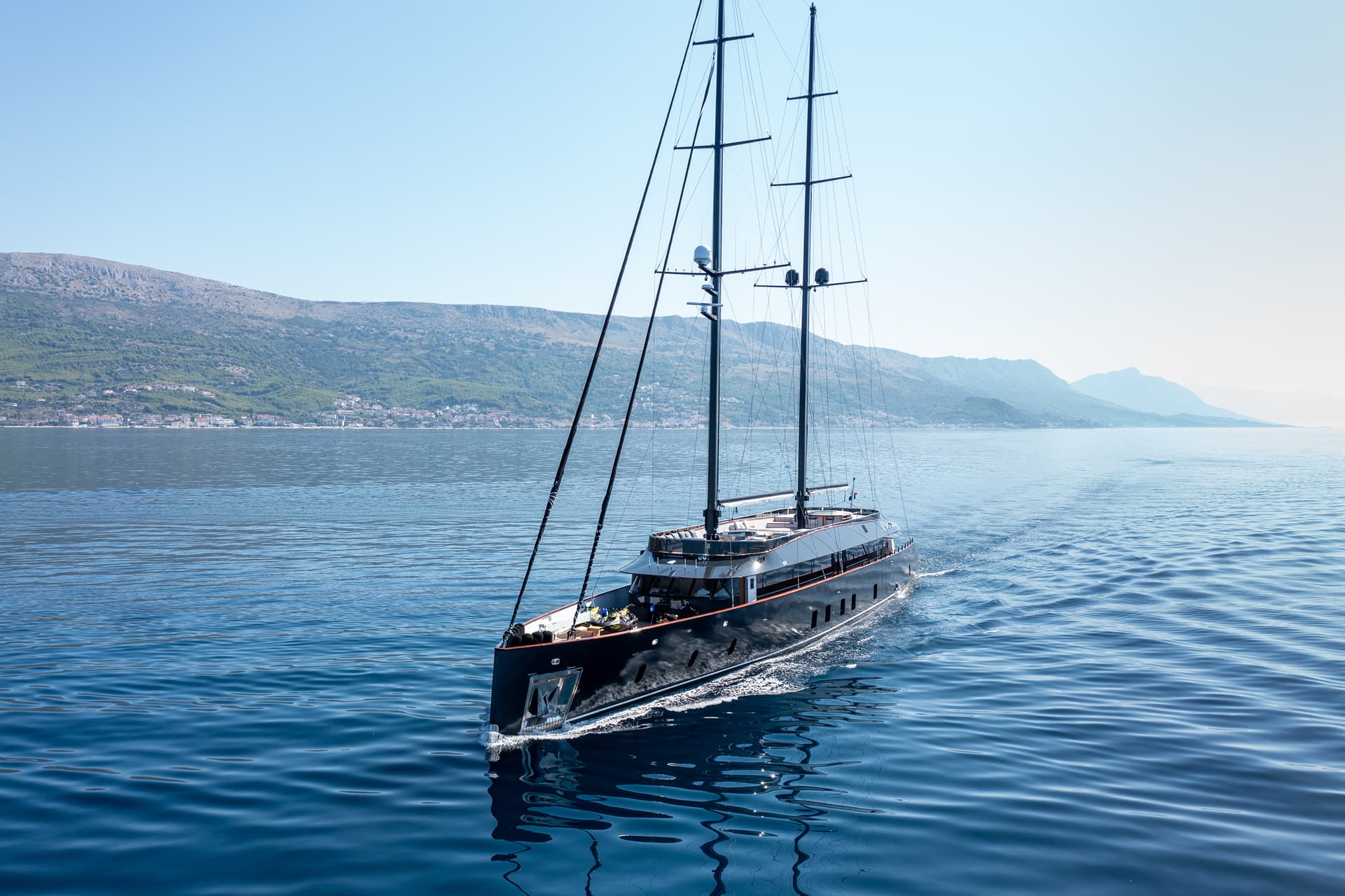 Large black sailing yacht cruising on blue water near a coastal mountain town.