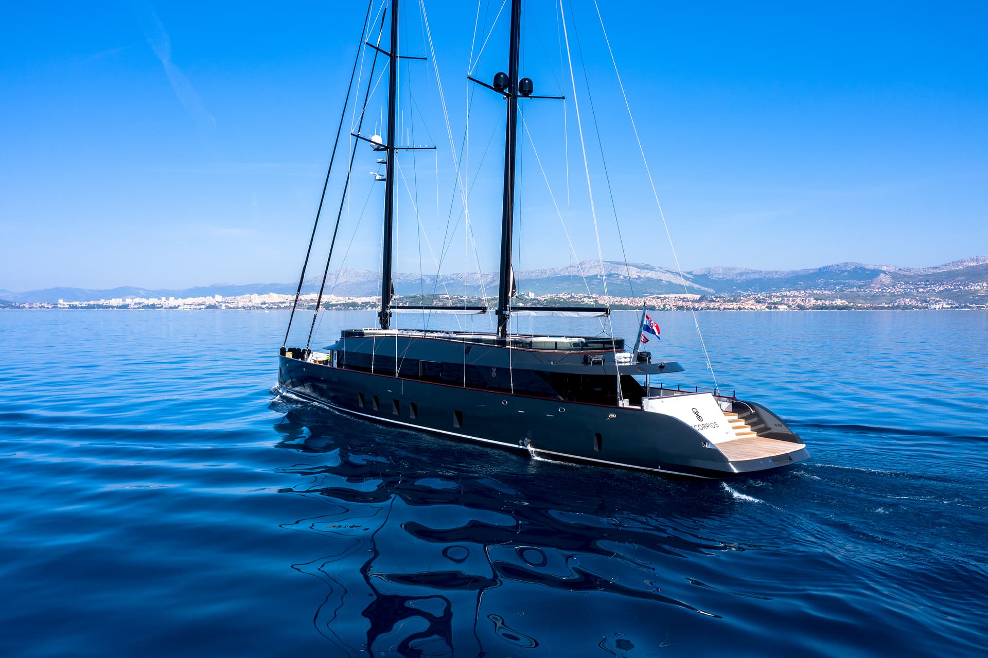 Large black sailing yacht named Scorpios cruising on deep blue water near a coastal city.