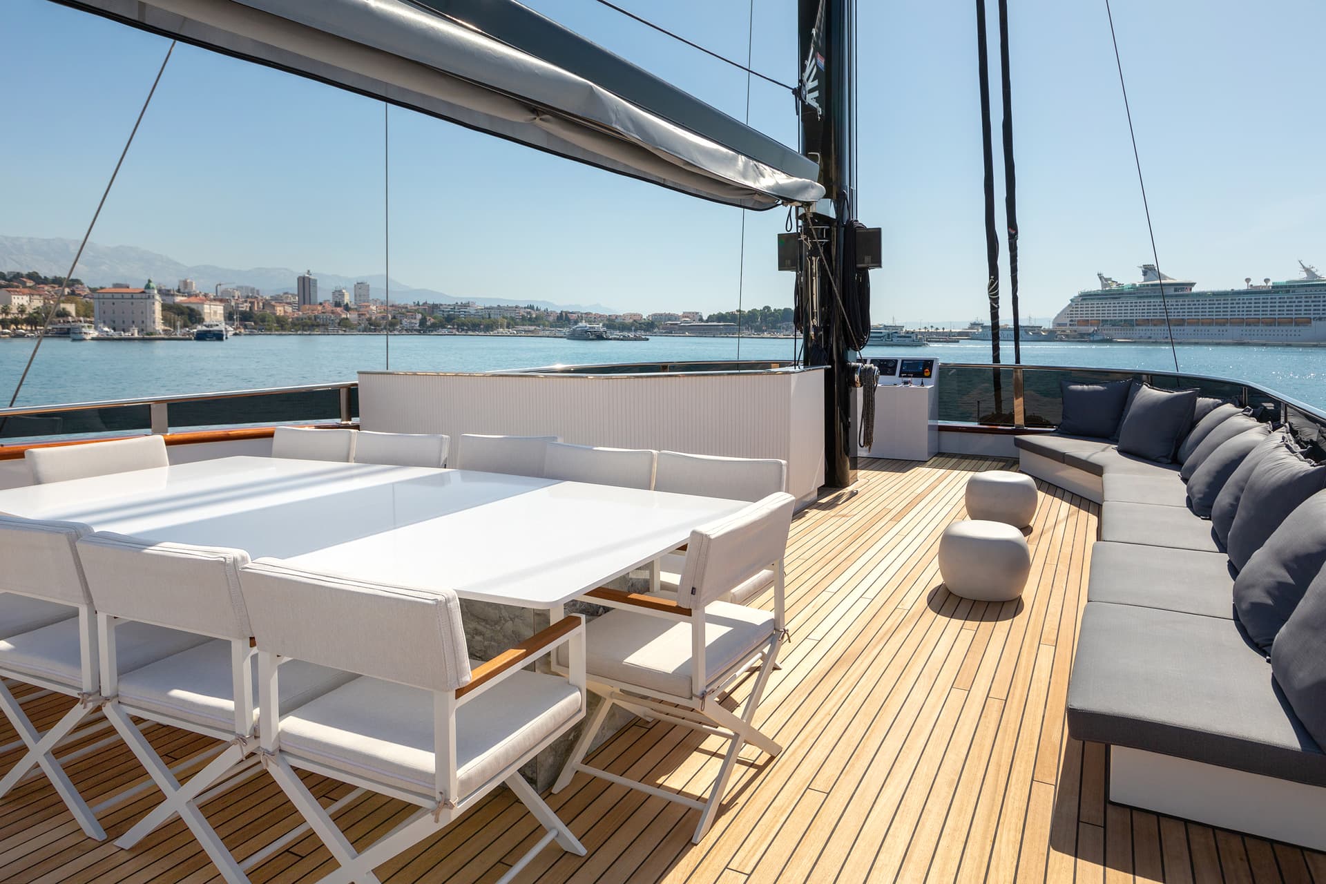 Yacht deck with white dining set and grey cushions overlooking a coastal city and cruise ship.