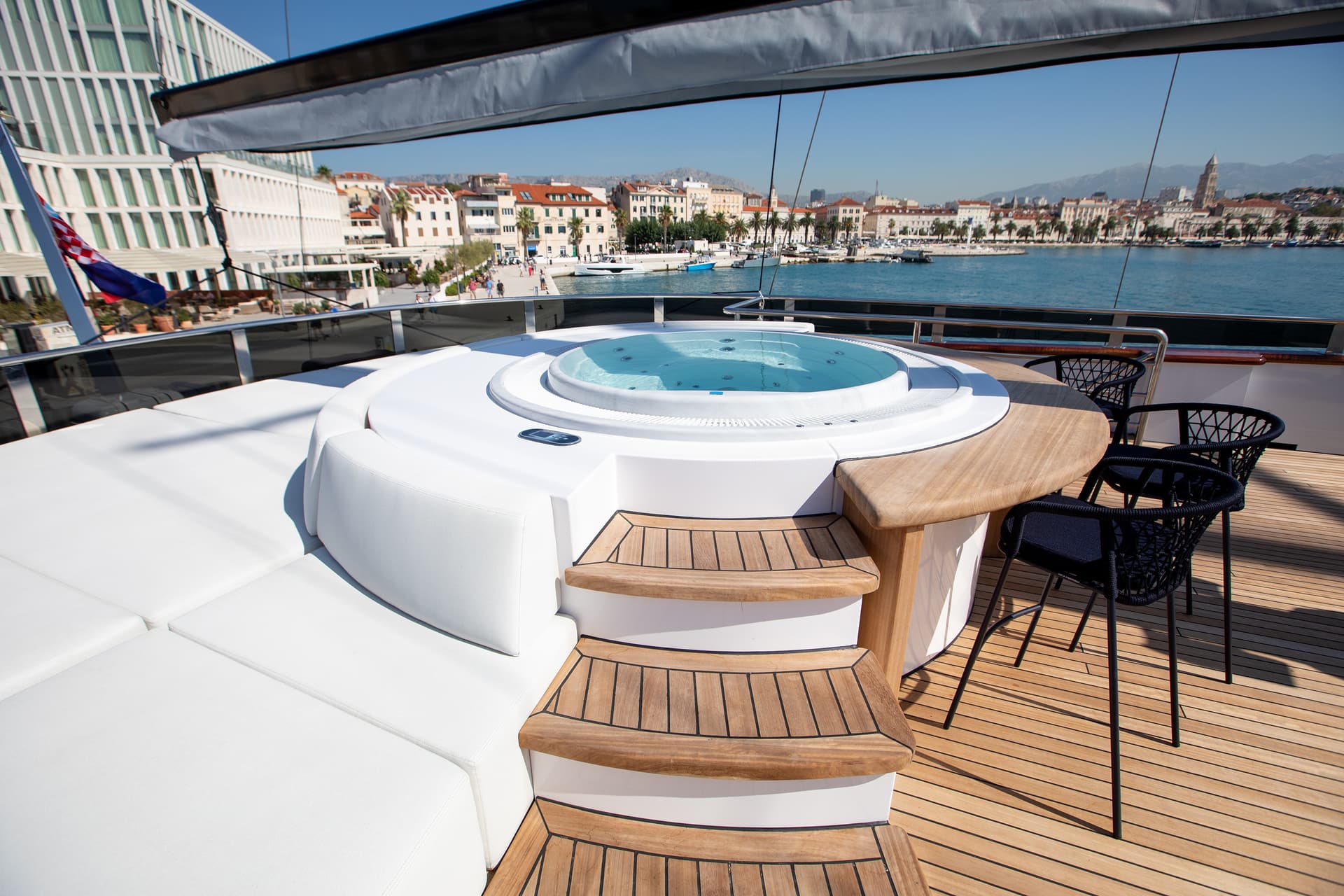 Yacht deck with hot tub and white seating overlooking coastal city and mountains.