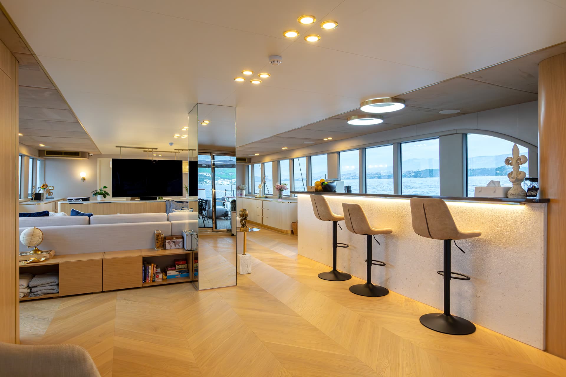 Modern yacht interior lounge with bar, light wood floors, and sea view of coastline.