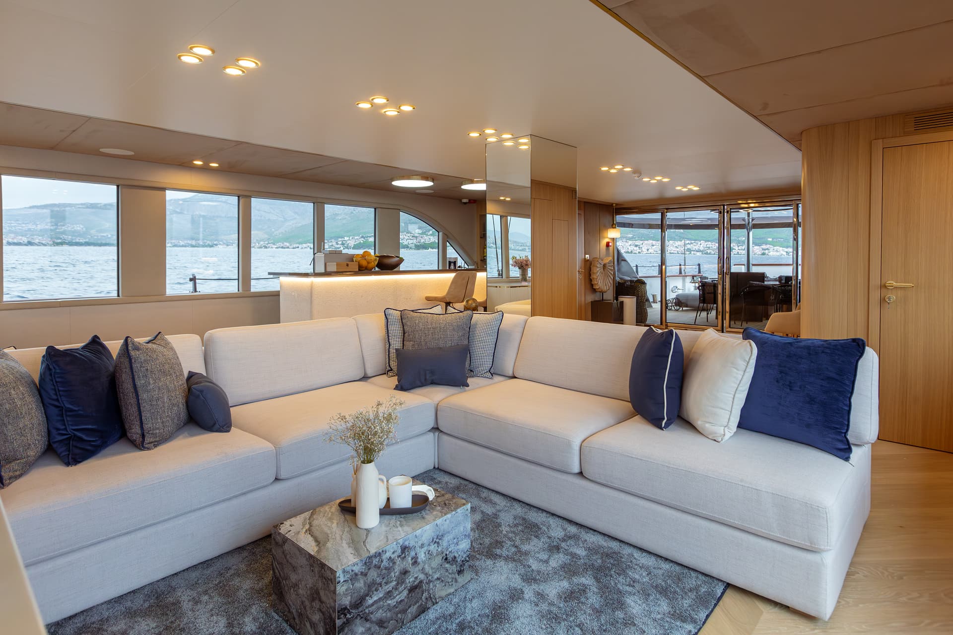 Yacht salon with light sofa, marble table, and coastal mountain view through windows.