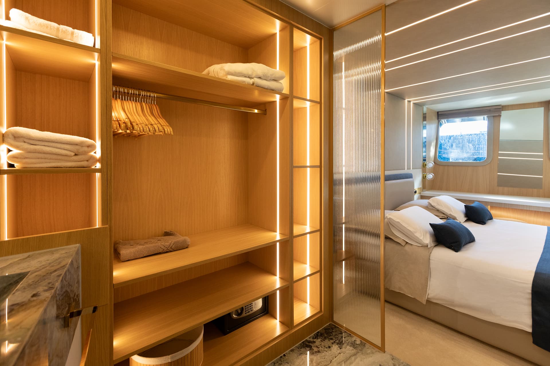 Luxury yacht cabin interior with illuminated wood shelving, safe, and bed near a porthole window.