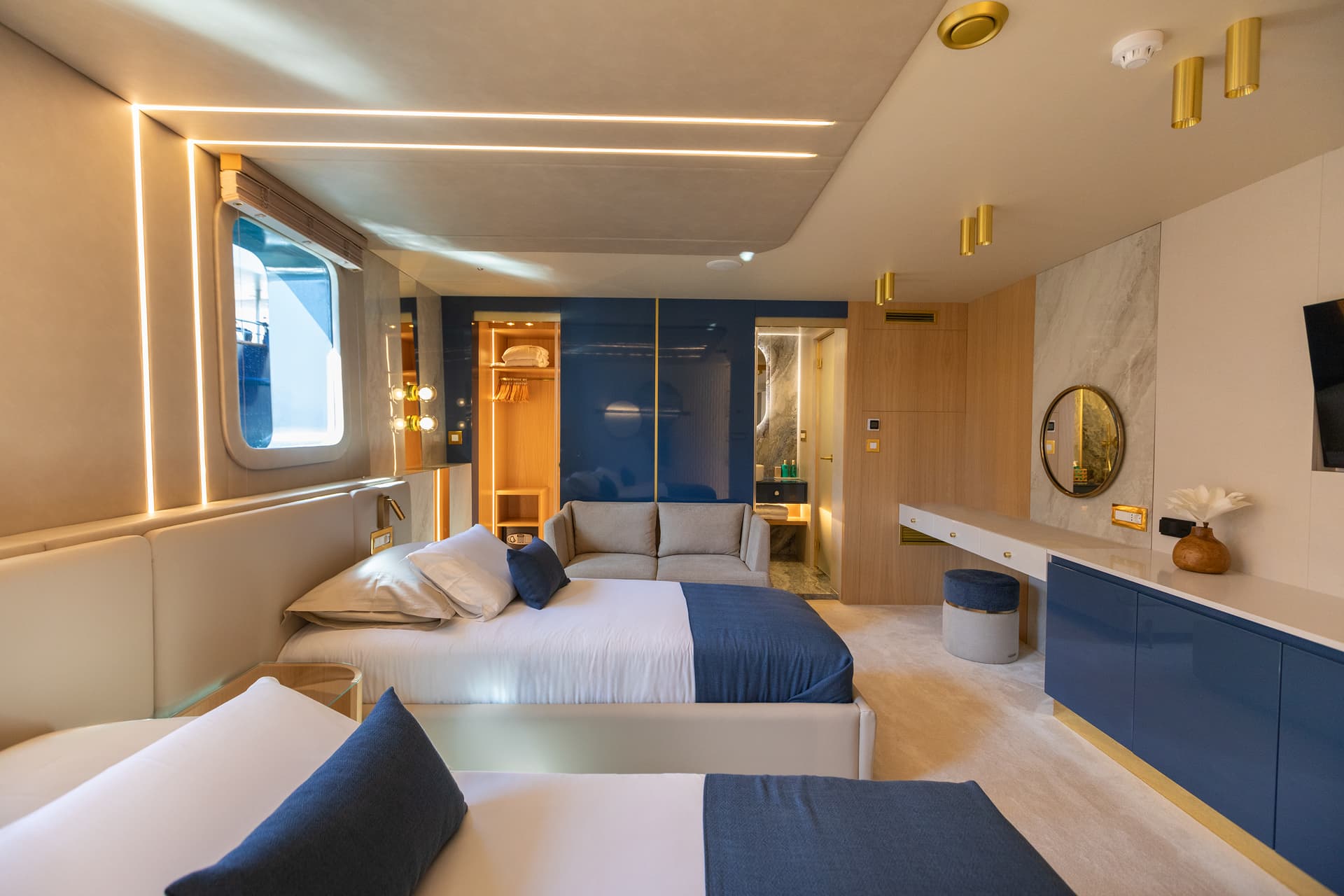 Modern yacht cabin interior with twin beds, blue accents, and ambient lighting near a porthole window.