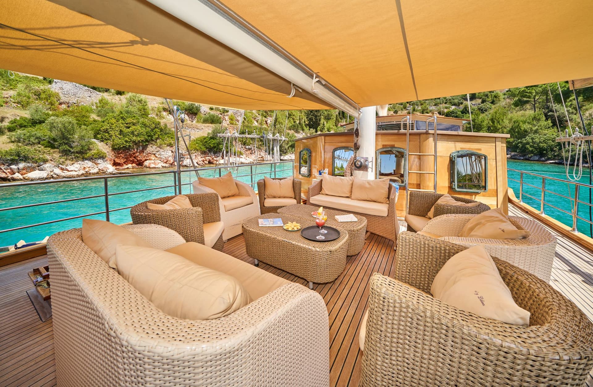 Wicker outdoor seating area on a yacht deck near a lush, rocky coastline with turquoise water.