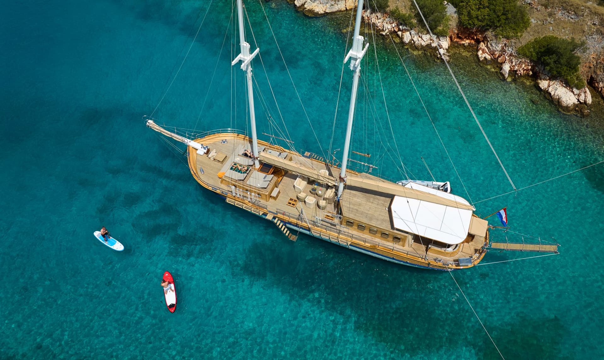 Large wooden sailboat anchored near rocky coast with two people paddleboarding in turquoise water.