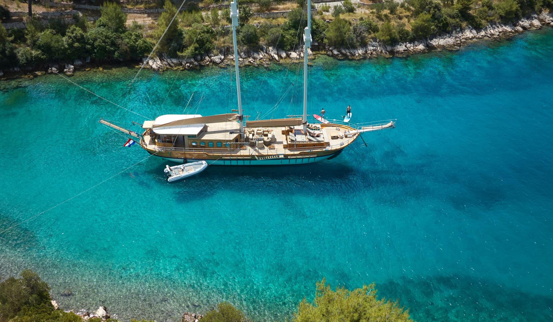 Large wooden sailboat anchored in turquoise coastal waters near a rocky, green shoreline.