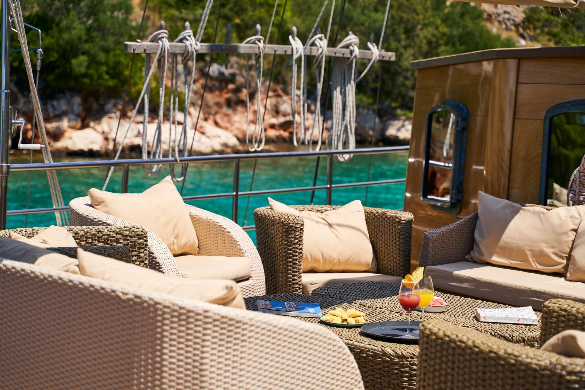 Wicker outdoor seating area on a boat deck overlooking turquoise water and a green coastline.