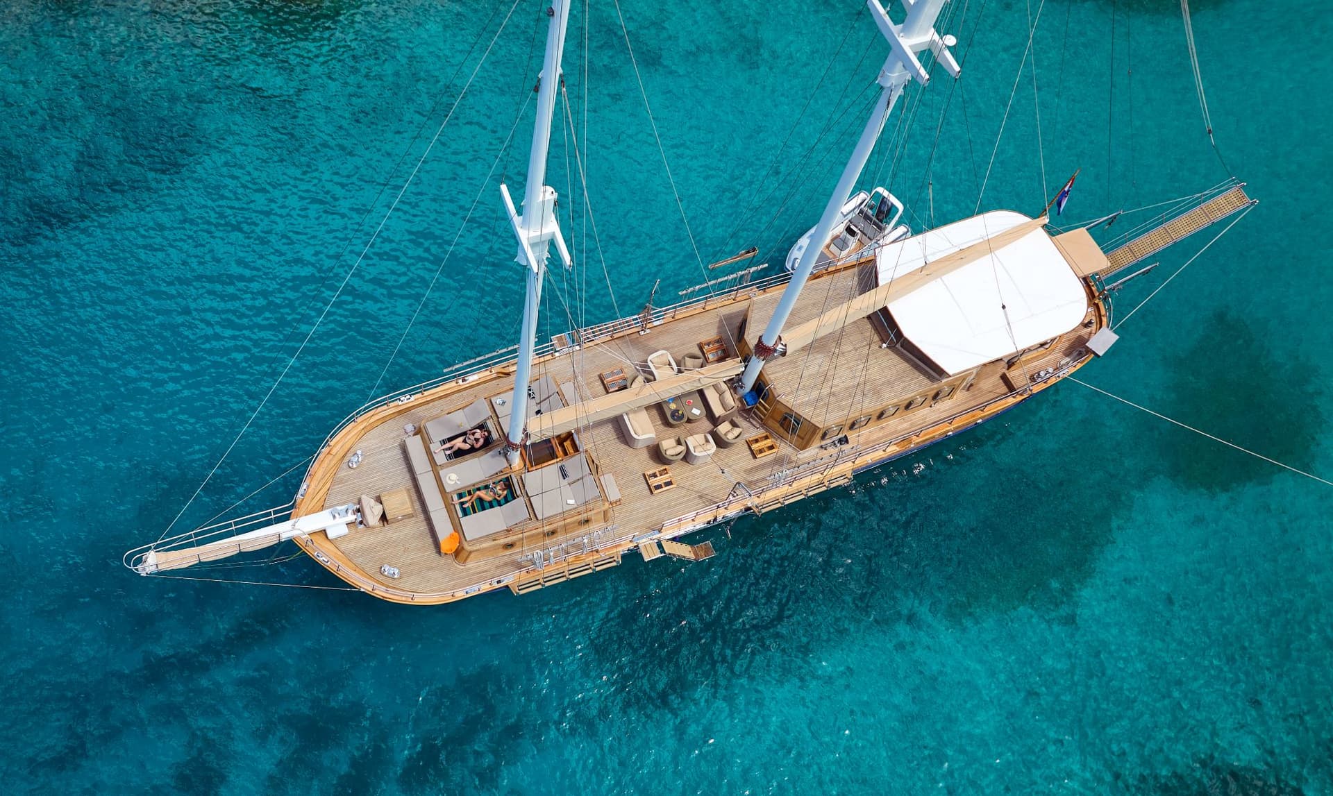 Aerial view of wooden sailboat anchored in clear turquoise coastal waters with sunbathers on deck.