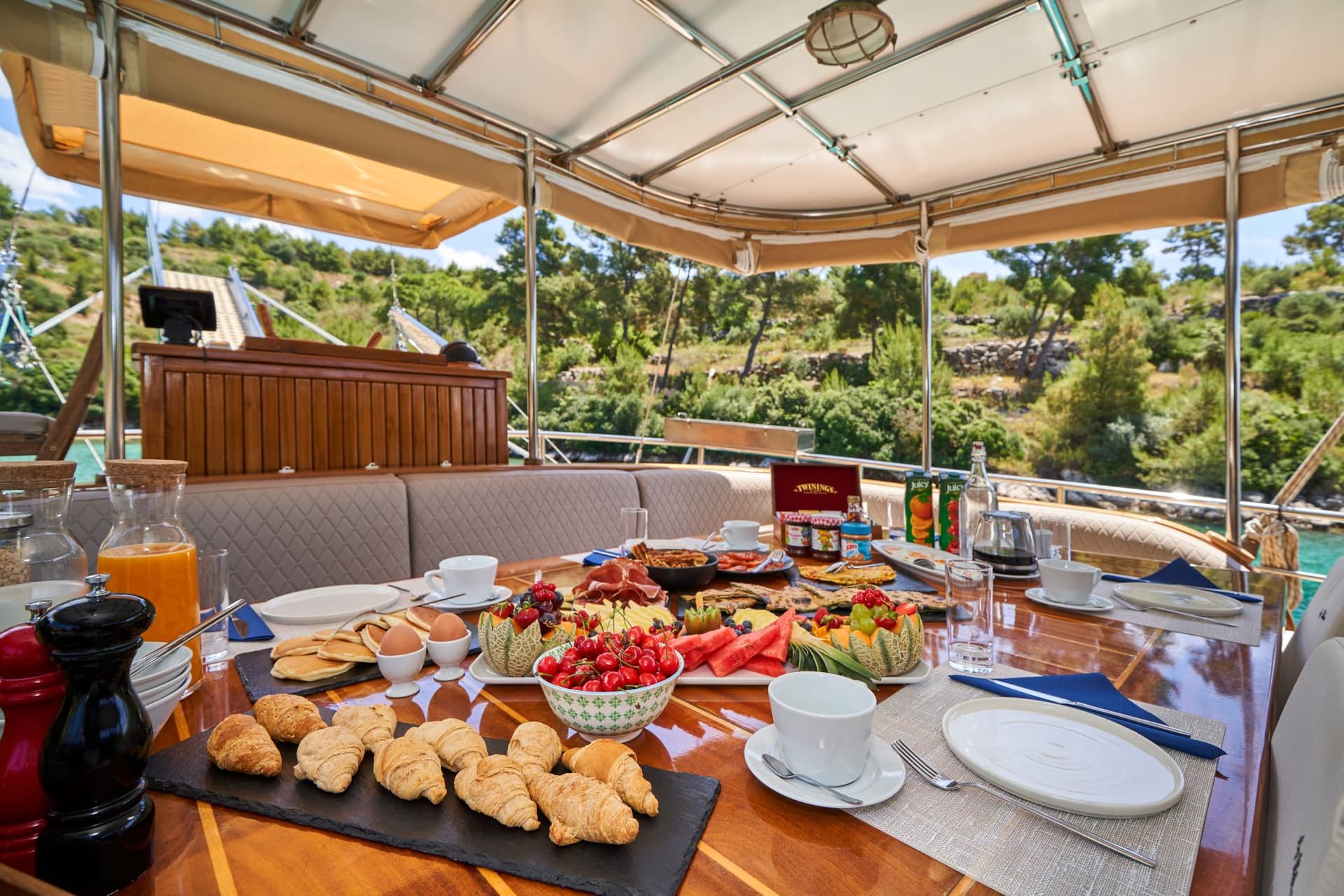 Breakfast spread with croissants, fruit, and coffee served on a boat deck near a wooded coastline.