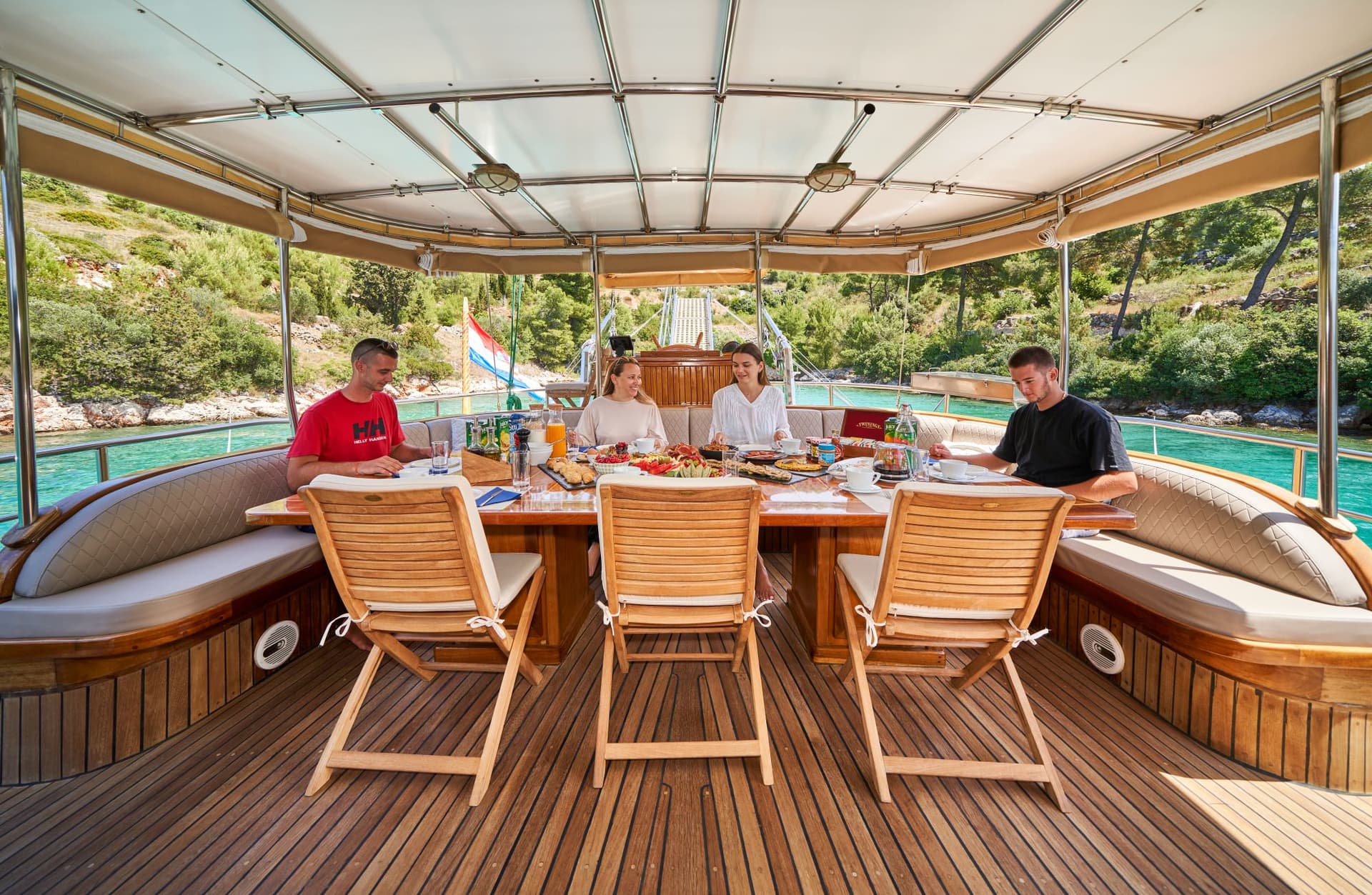 Group dining on boat deck with teak flooring near lush green coastline and turquoise water