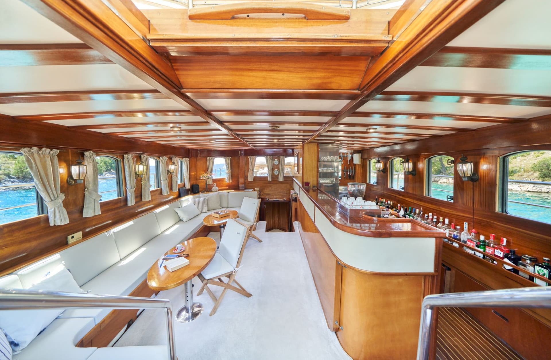 Interior saloon of a wooden boat with a bar, white seating, and views of turquoise water.