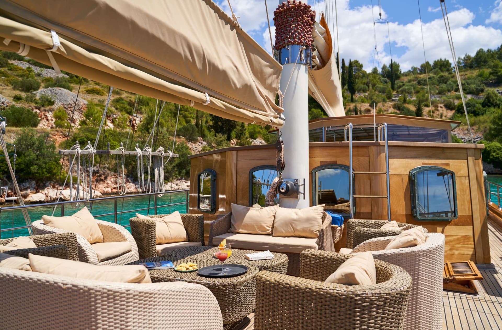 Wicker lounge area on a boat deck near a lush, green, rocky coastline.