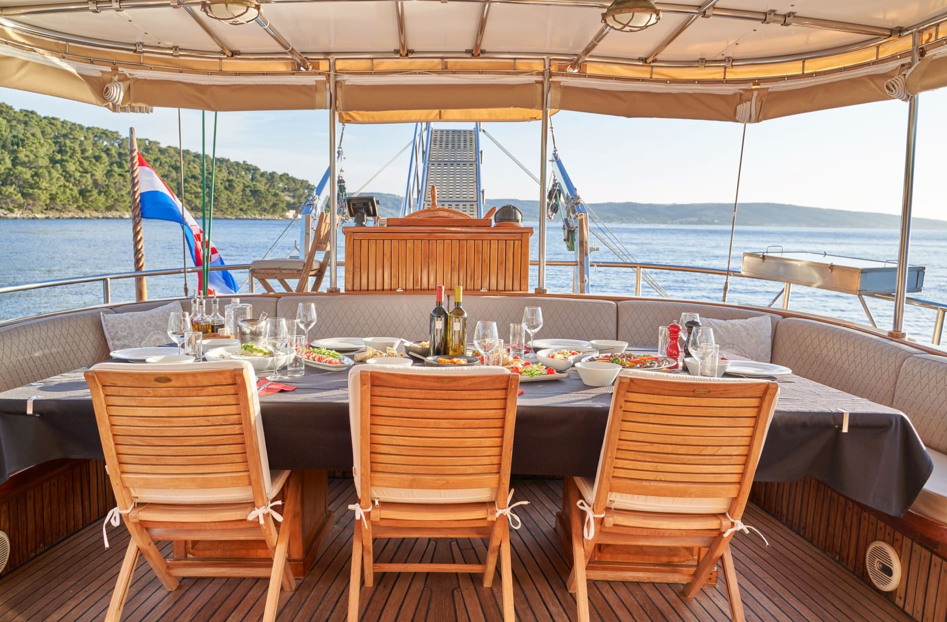 Outdoor dining area set on a yacht deck near a forested coastline.